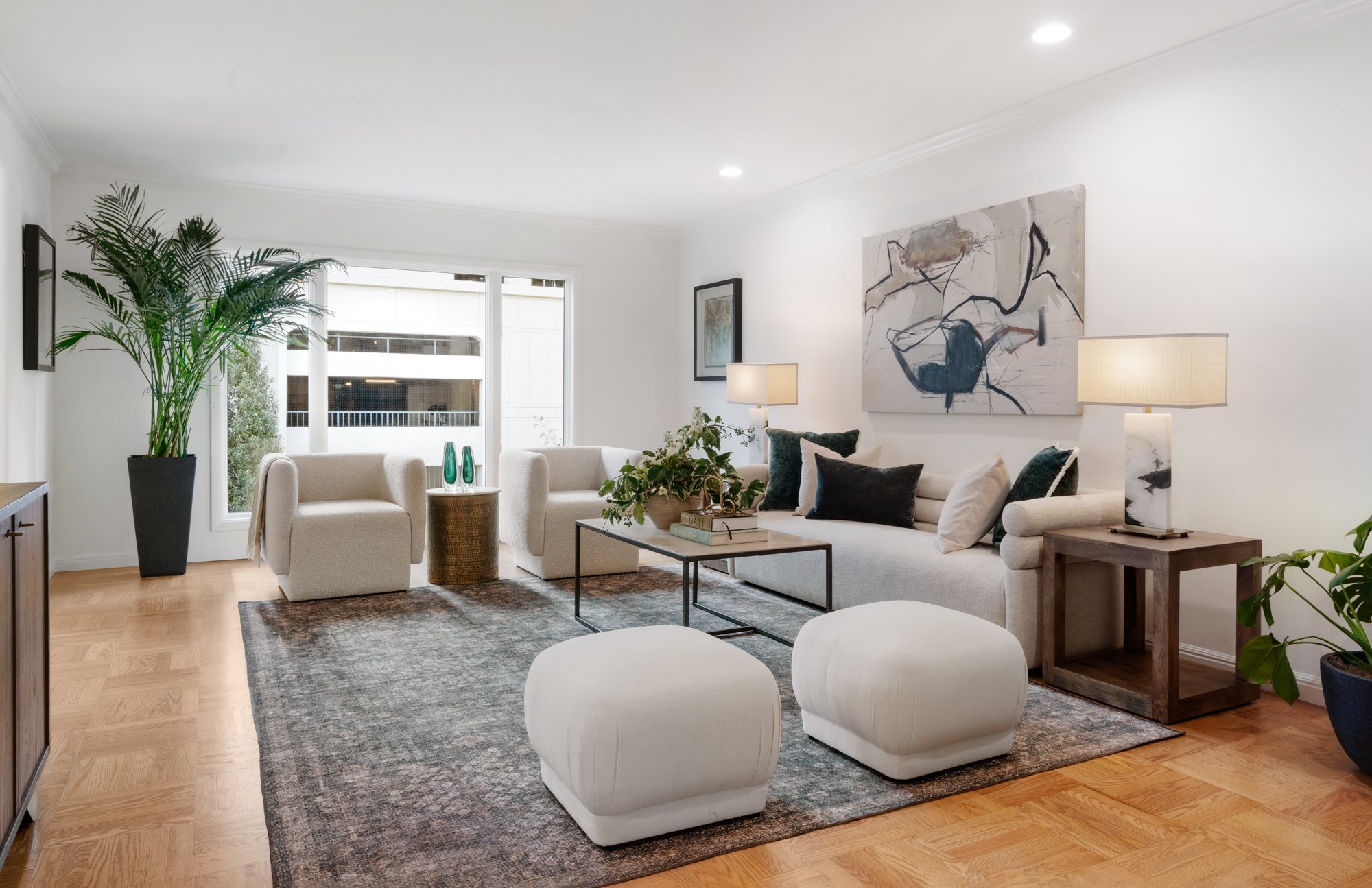 Bright living room with white sofa, two white ottomans, two armchairs, side tables with lamps, large window, abstract art on wall, houseplants, and a gray area rug.