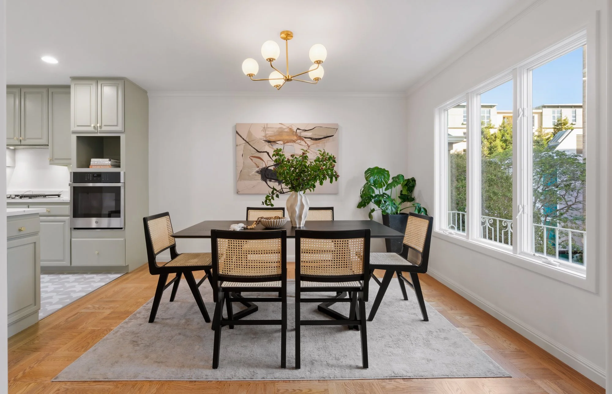Dining room with six black and rattan chairs around a dark wooden table, a large white watercolor vase with green plants, amid wall art and a large window showing trees outside.