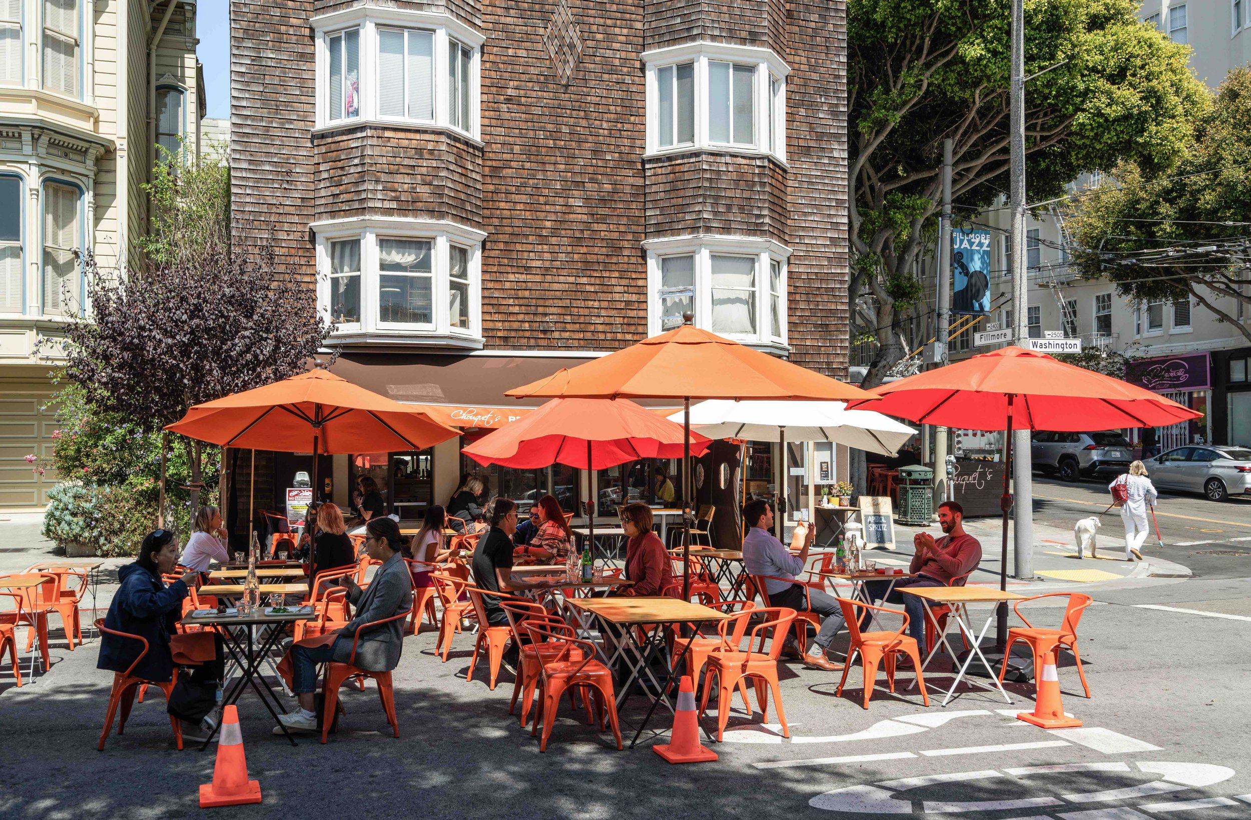 Outdoor cafe with orange umbrellas and seating, people dining, busy street scene on a sunny day in an urban neighborhood with historic apartment buildings and trees.