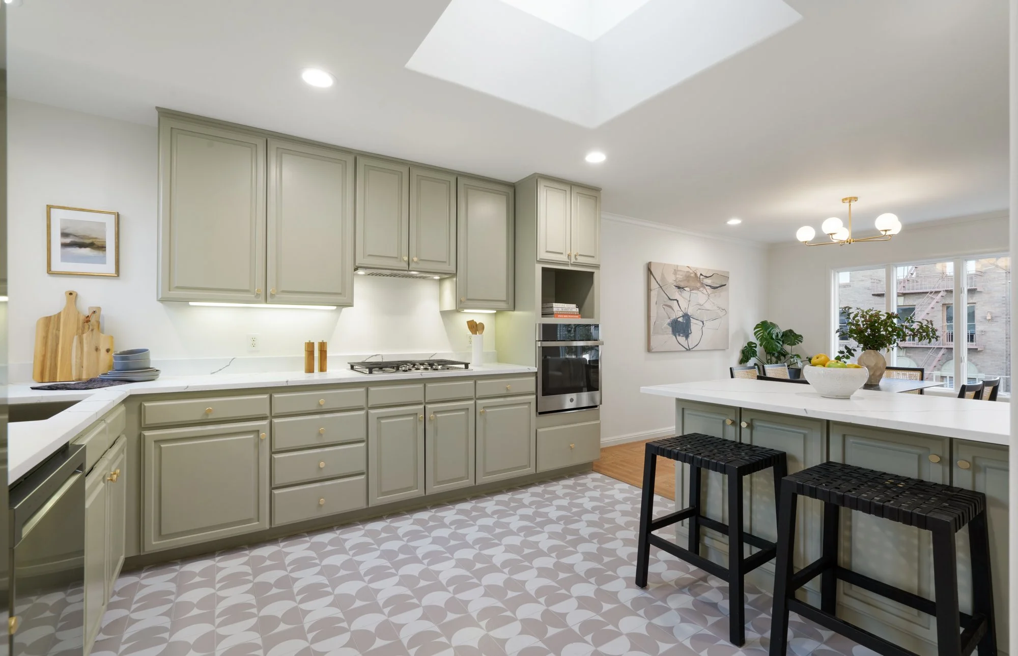 Modern kitchen with green cabinets, white countertops, and patterned floor tiles. Features include an oven, stovetop, wooden cutting boards, and a breakfast bar with two black stools. In the background, there is a dining area with a table, chairs, an