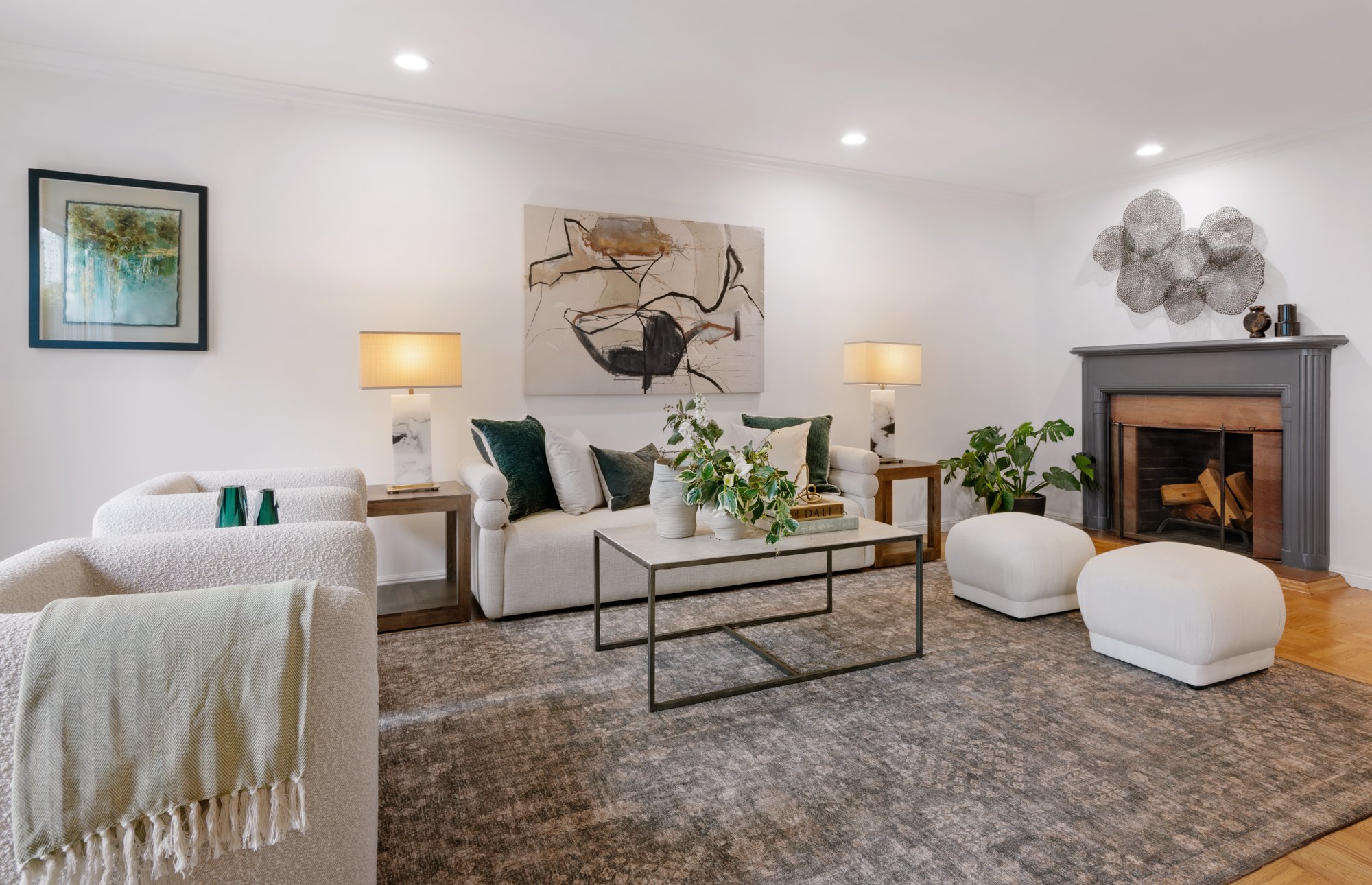 A modern living room with white walls, a gray fireplace, abstract artwork, a white sofa with dark green and cream pillows, a glass top coffee table with decorative vases and greenery, two cream ottomans, wall-mounted lights, a framed landscape painting, and a textured area rug.