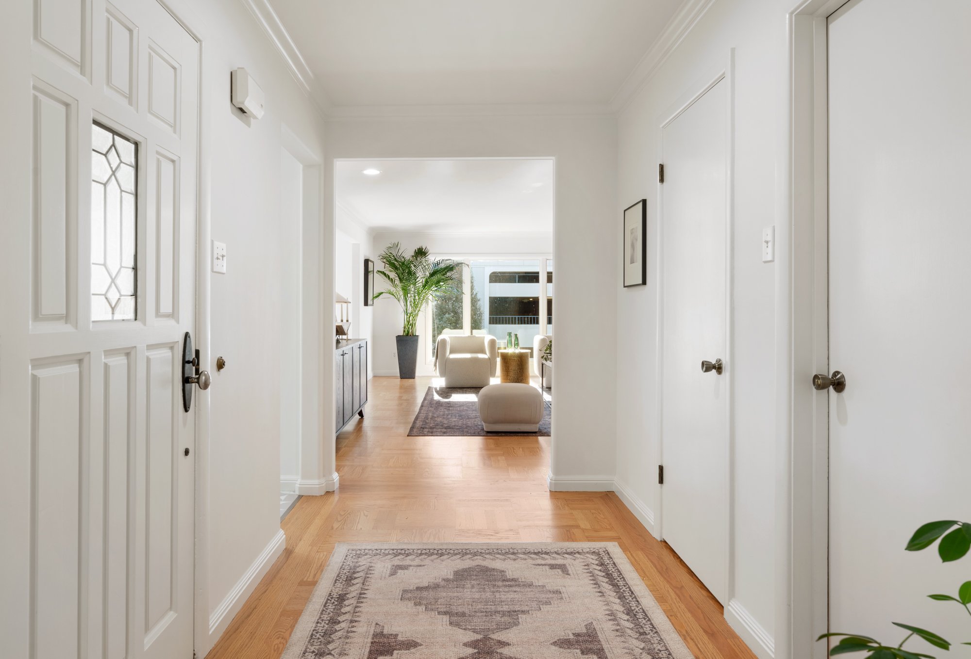 Clean and bright hallway leading into a living room with large window, potted plant, and modern furniture.