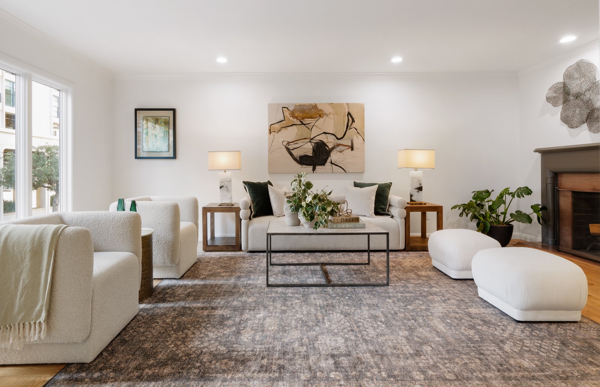 Living room with white walls, large window, modern white sofa, abstract artwork, two side tables with lamps, green plants, and contemporary decor.