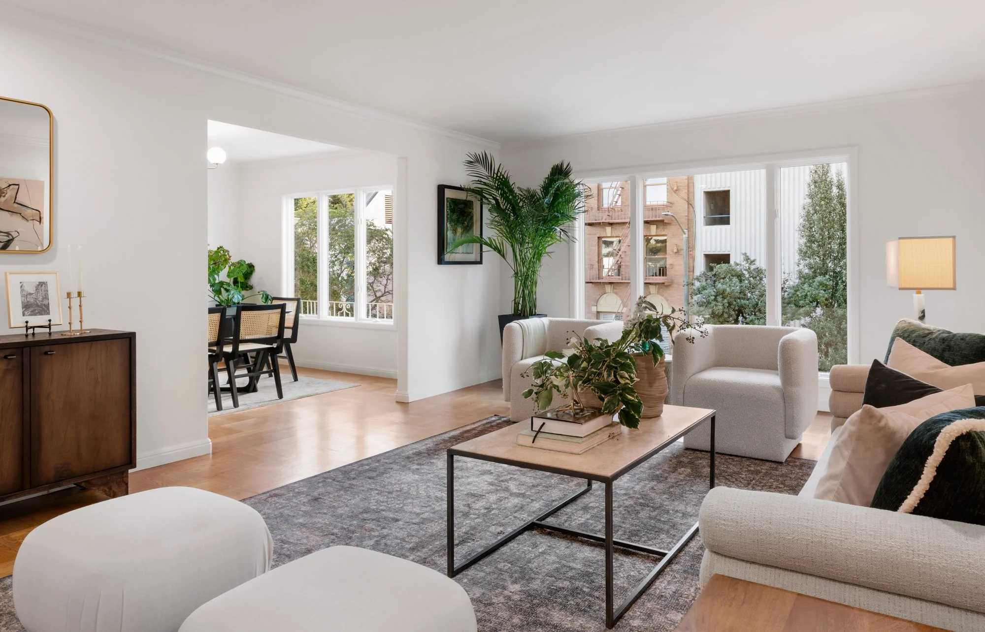 Bright living room with large windows, white sofas, a wooden coffee table with plants, and a view of neighboring buildings and trees.