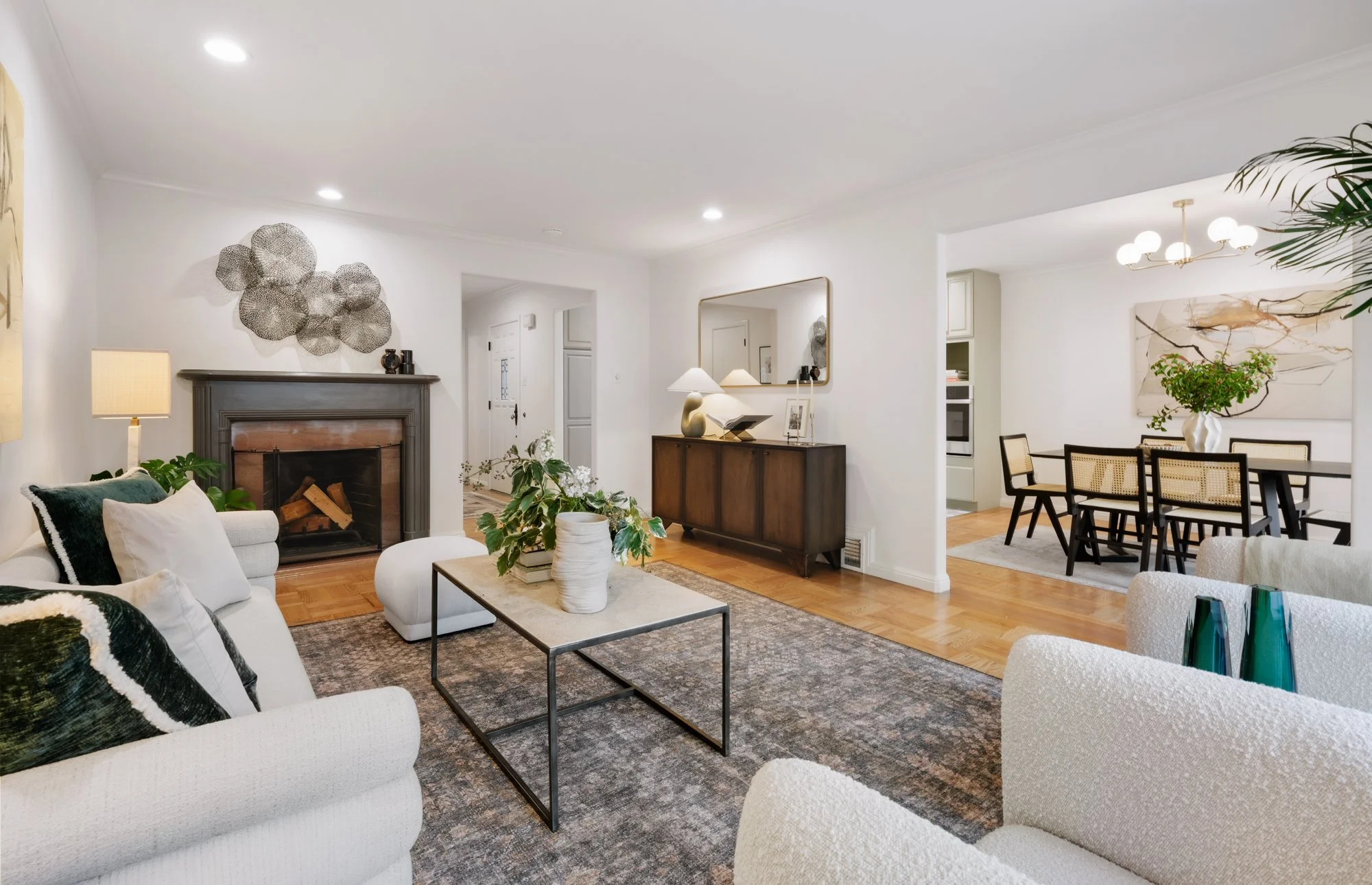 Modern living room with white walls, hardwood floors, a fireplace, white sofa, black and white pillows, a coffee table with vases and a plant, framed artwork, a sideboard with a lamp, mirror, and decorations, and a dining area with a table, chairs, a