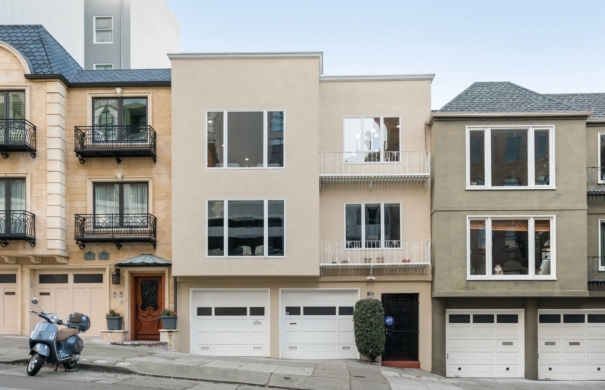 Front view of three adjacent modern multi-story residential buildings with balconies, large windows, and garage doors, with a scooter parked on the street in front.