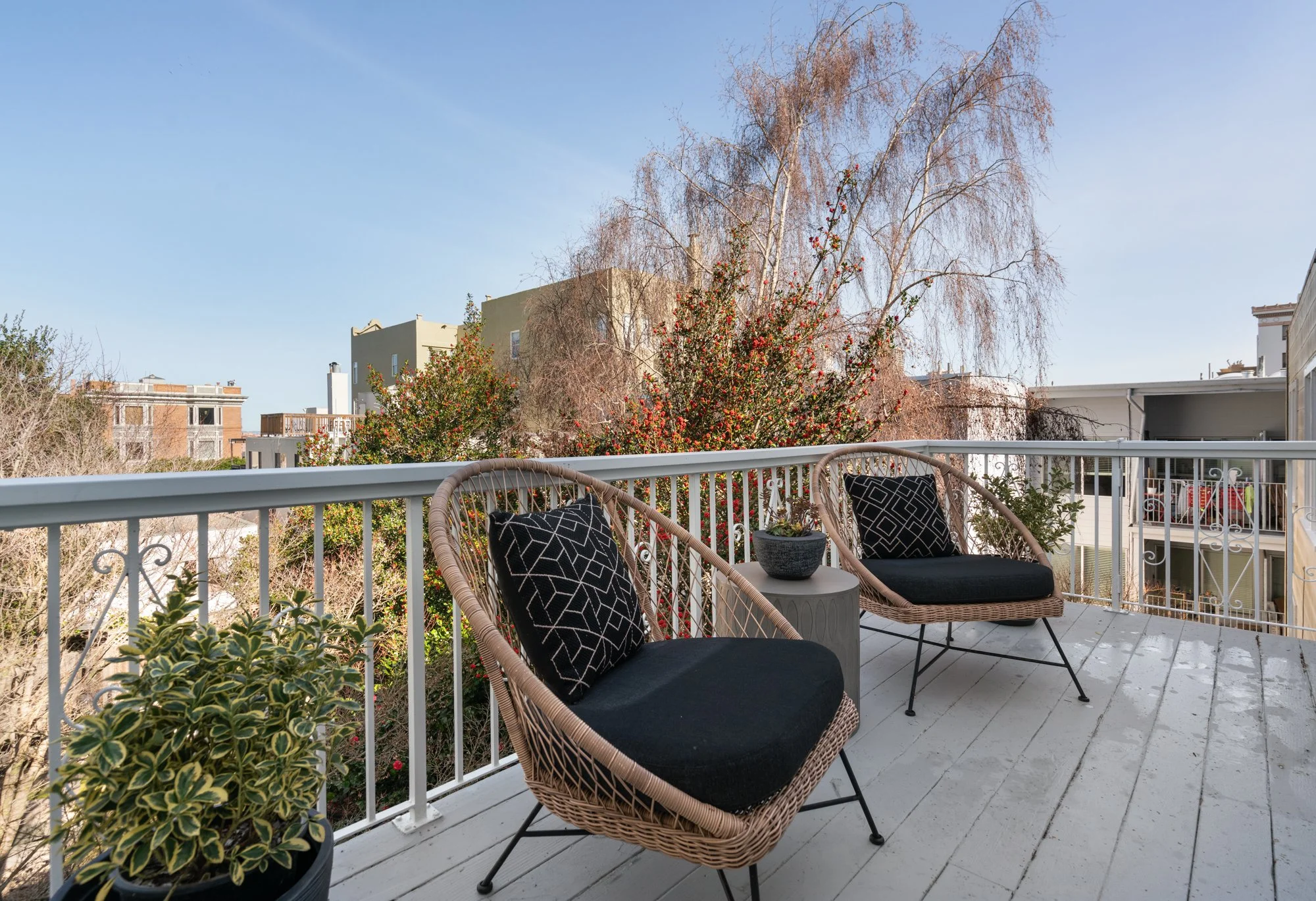 Balcony with two wicker chairs with black cushions and patterned pillows, a small round table with a potted plant, overlooking neighboring buildings and trees, under a clear blue sky.