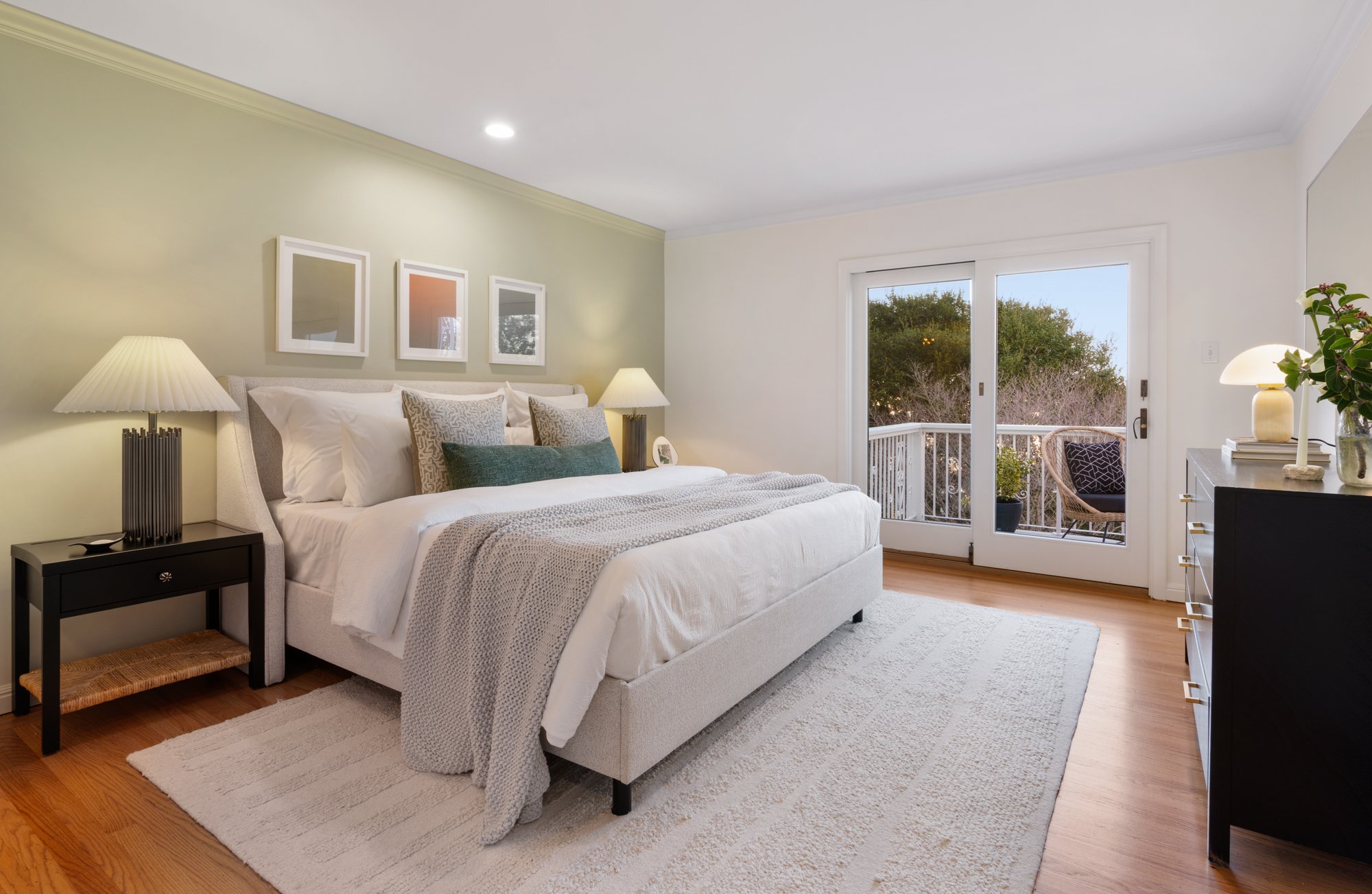 A bright bedroom with a white bed, beige headboard, and decorative pillows. Nightstands with lamps on each side, a black dresser with a lamp and plant, and a sliding glass door leading to a balcony with outdoor seating. Light-colored walls and hardwood flooring.