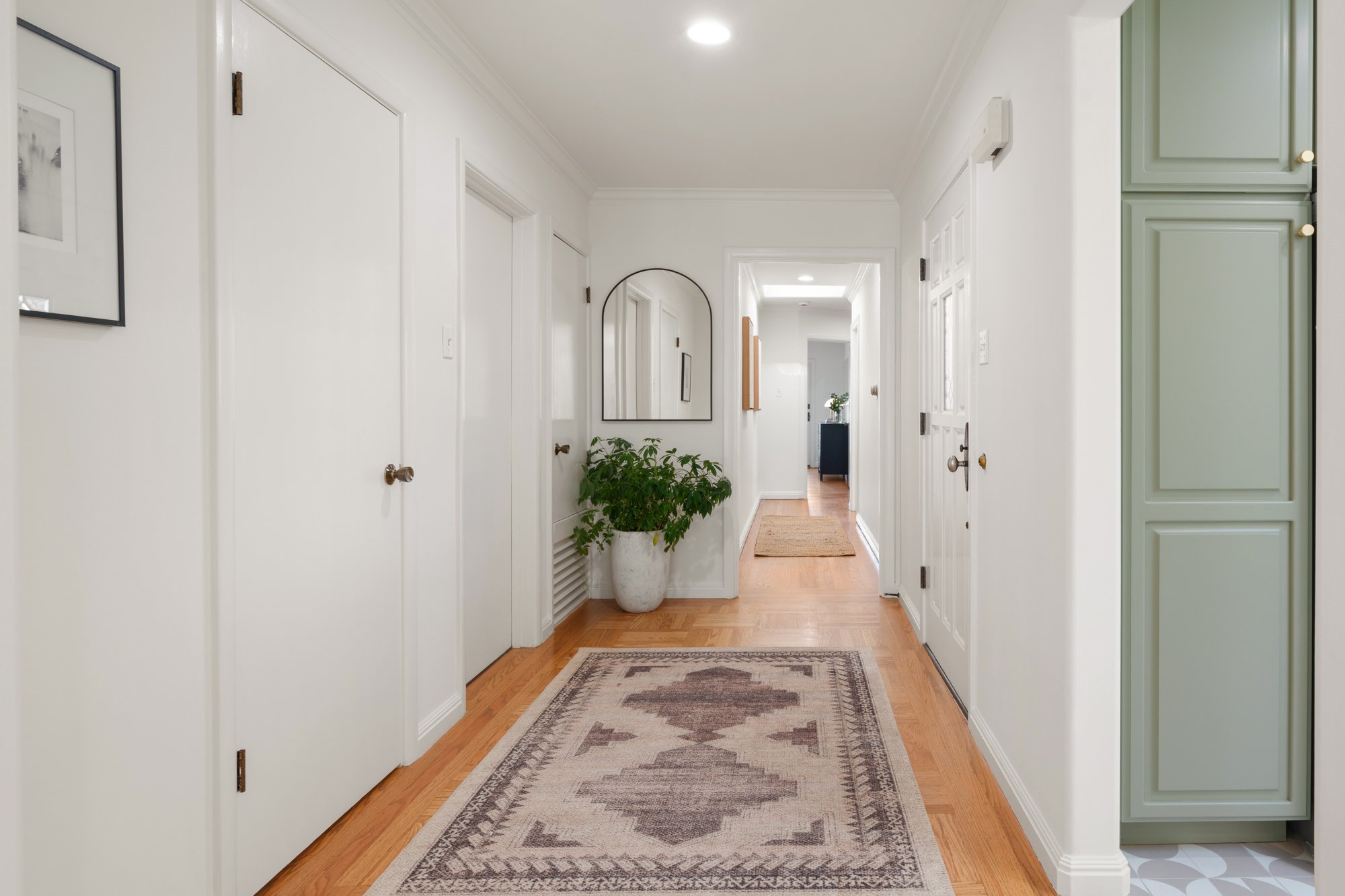 Bright hallway with white walls, wooden flooring, a green cabinet on the right, a large potted plant on the left, a rectangular mirror on the wall, and a patterned rug on the floor.