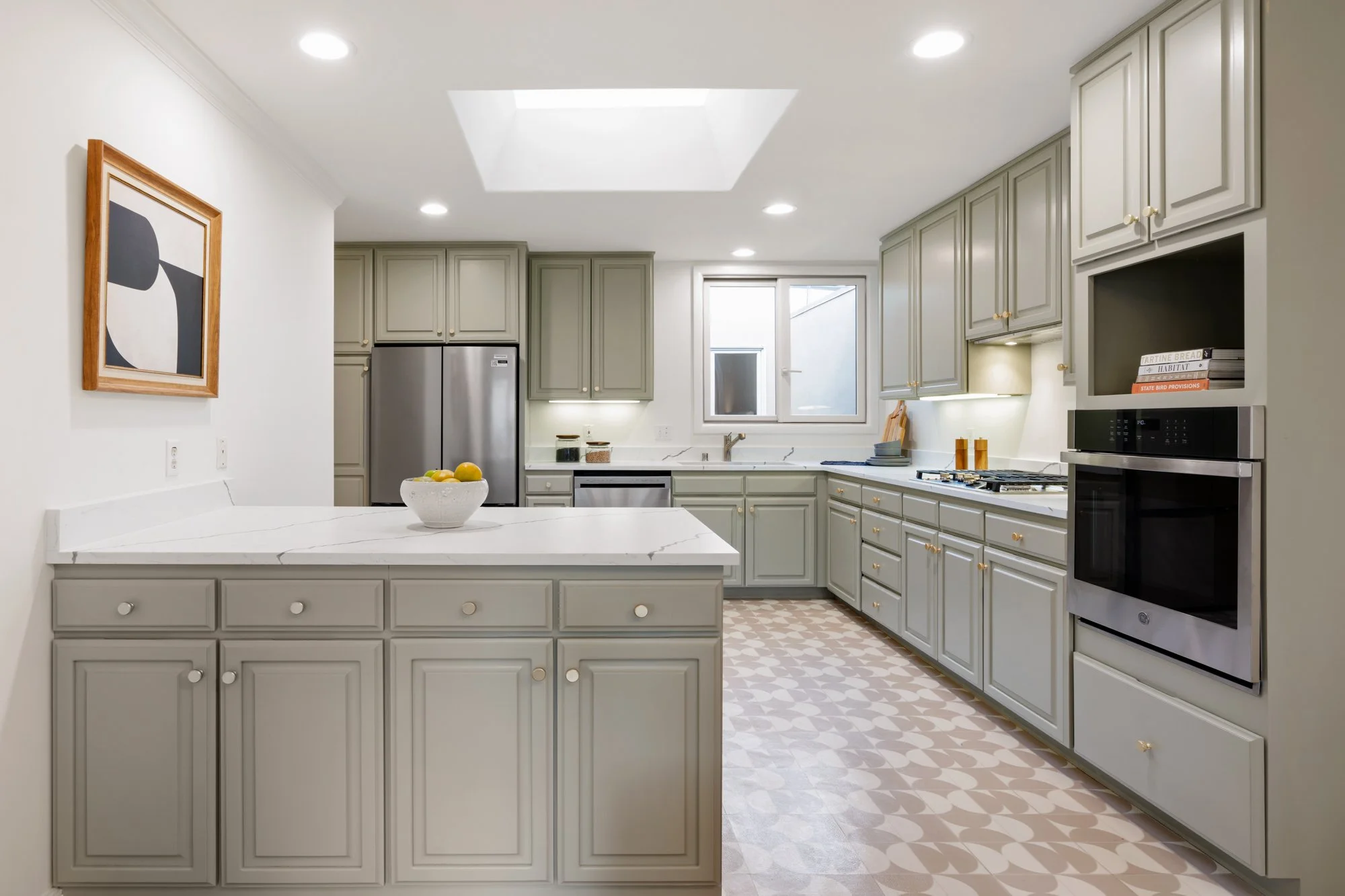 Modern kitchen with light gray cabinets, white marble countertops, stainless steel appliances, a bowl of lemons on the island, patterned floor tiles, a window, and a framed abstract art piece on the wall.