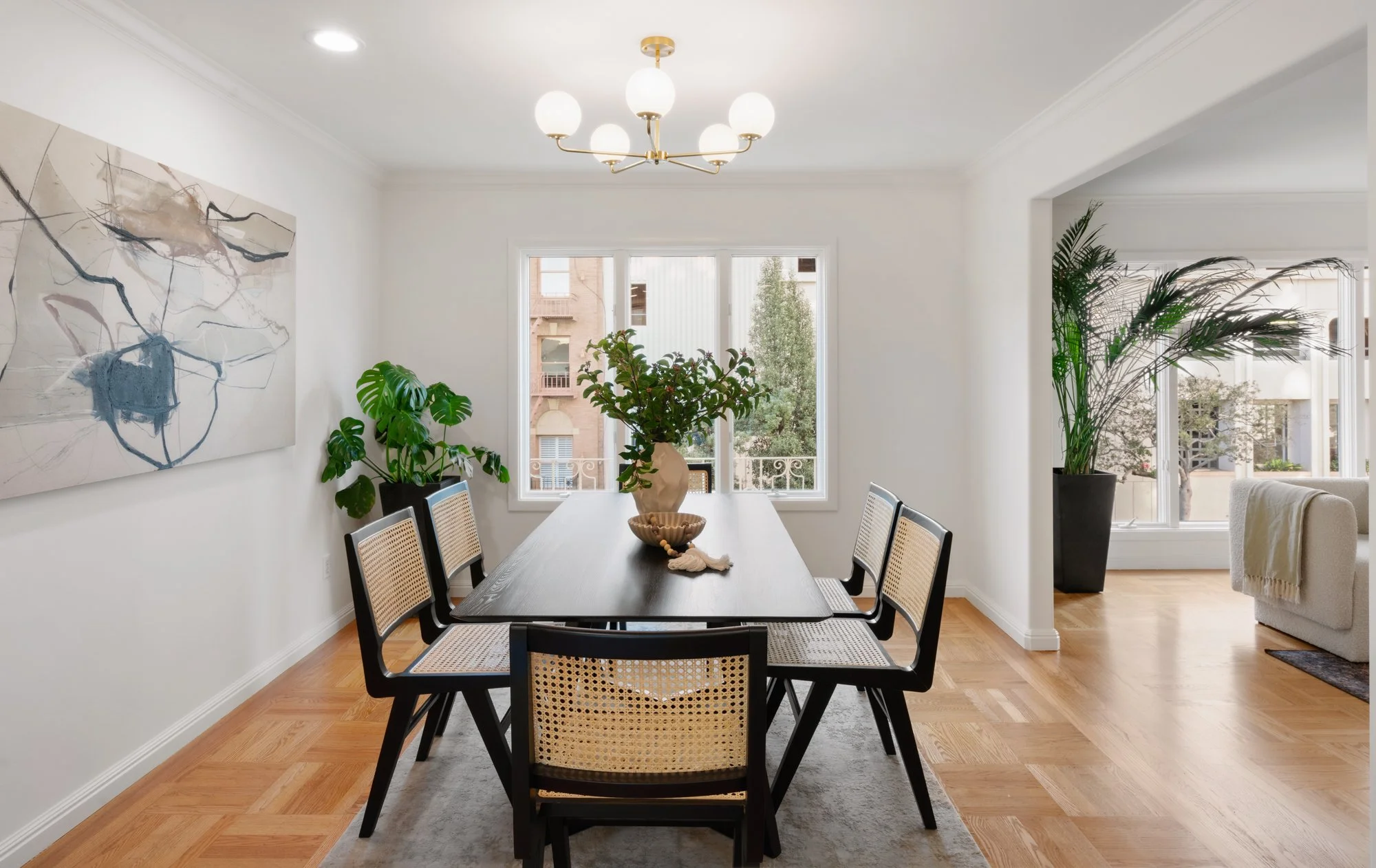 Dining room with a dark wooden table, six chairs, a large window, indoor plants, a piece of abstract artwork on the wall, a chandelier on the ceiling, and hardwood flooring.