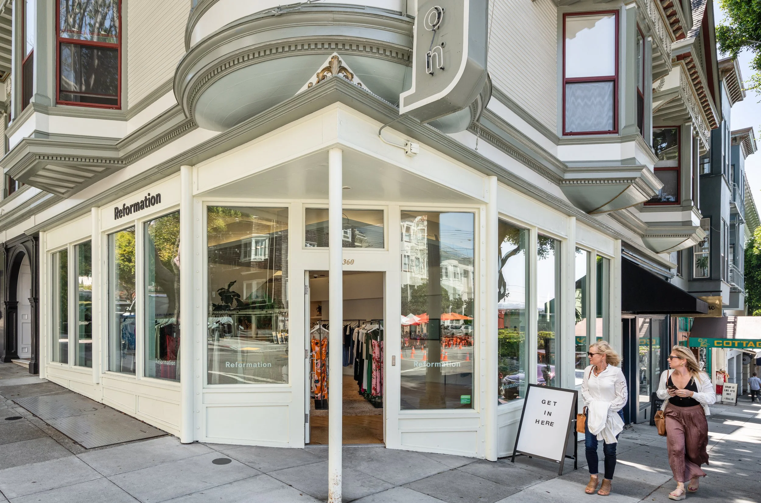 A corner storefront with large windows displaying clothing, a sign that reads 'GET IN HERE,' and two women walking past on the sidewalk.