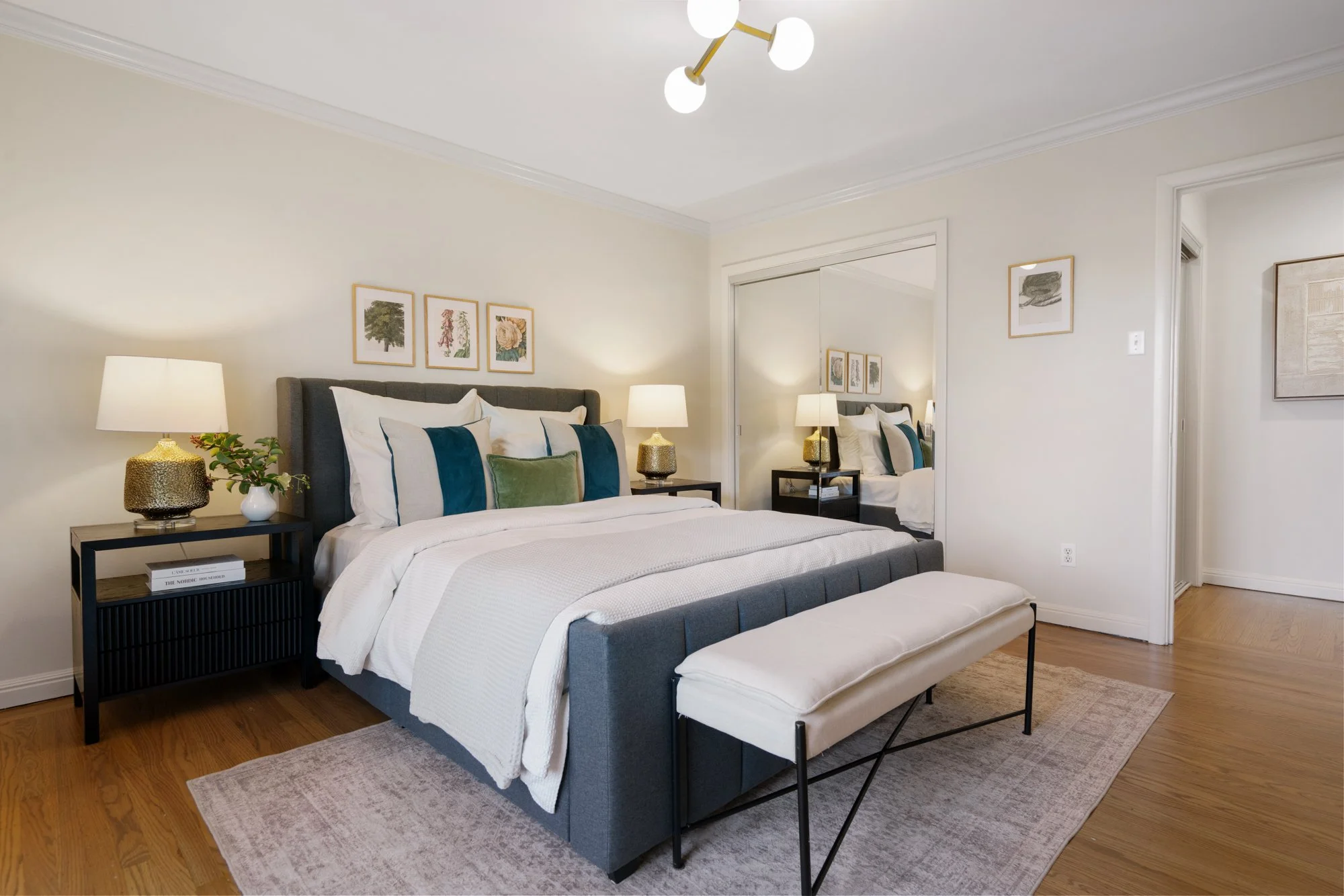 A bedroom with a gray upholstered bed, white bedding, and colorful pillows, flanked by black nightstands with lamps, a bench at the foot of the bed, and a large mirror reflecting part of the room.