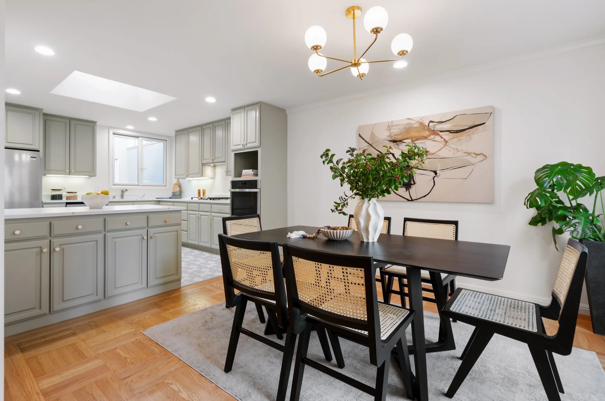 Dining area with a dark wooden table and six chairs, a large white vase with green foliage, spacious kitchen with light cabinets, a window, and modern lighting. Abstract art on the wall.