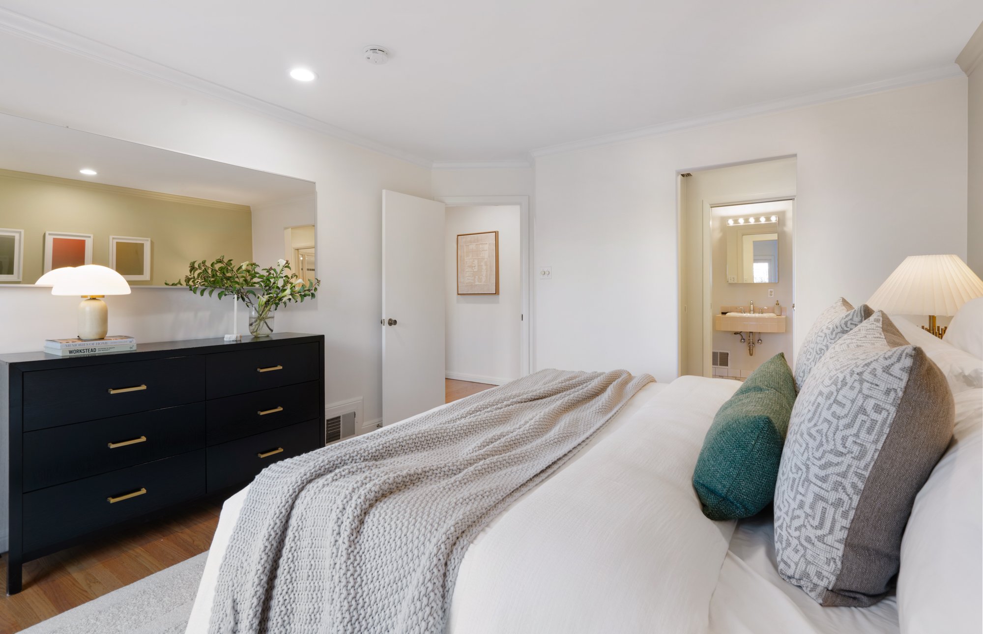 A cozy bedroom with a neatly made bed, decorative pillows, and a nightstand with a lamp. A dresser with a lamp, books, and a decorative plant is against the wall. There is a small bathroom with a sink visible through an open door.