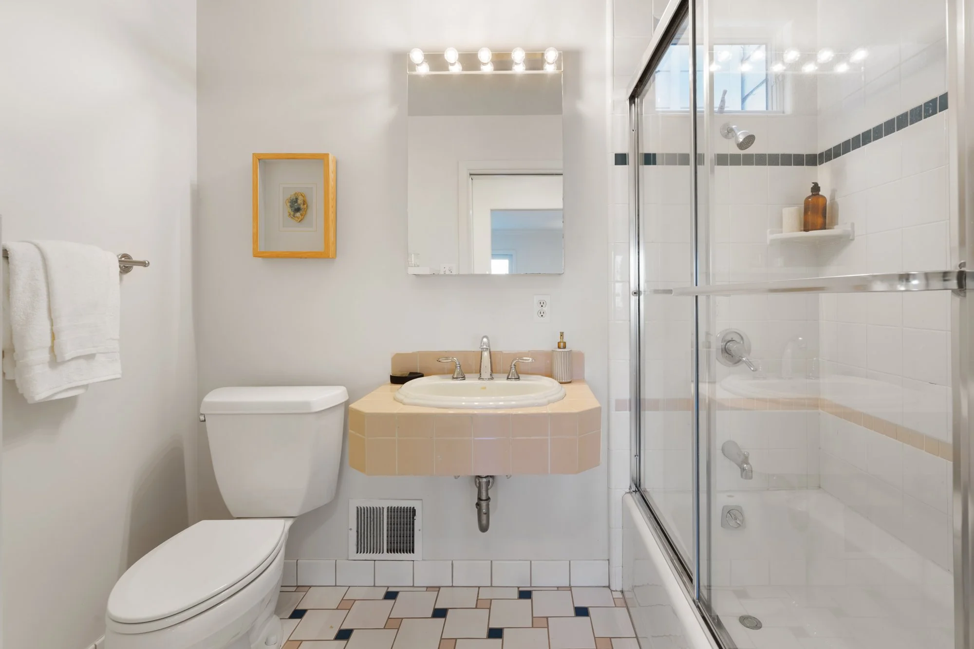 Bathroom with white toilet, beige sink with mirror above, glass shower with sliding door, small window, and white tiled walls.