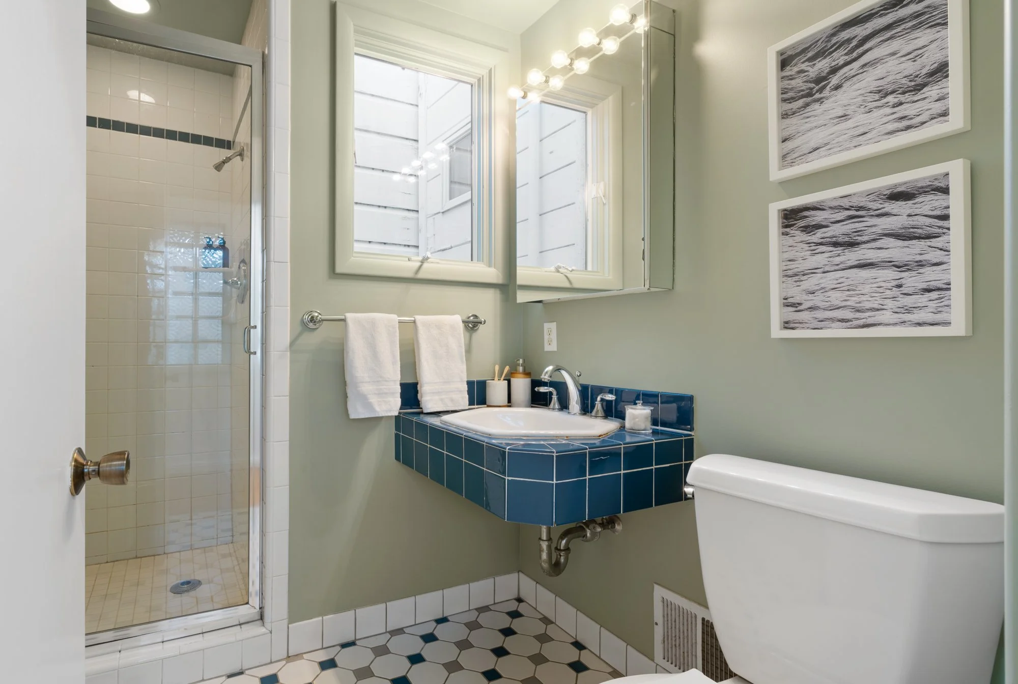 Bathroom with a glass-enclosed shower, a blue tiled sink with a white basin, a white toilet, two framed pictures of waves, green walls, a window, and a mirror. There are towels hanging on a towel bar.
