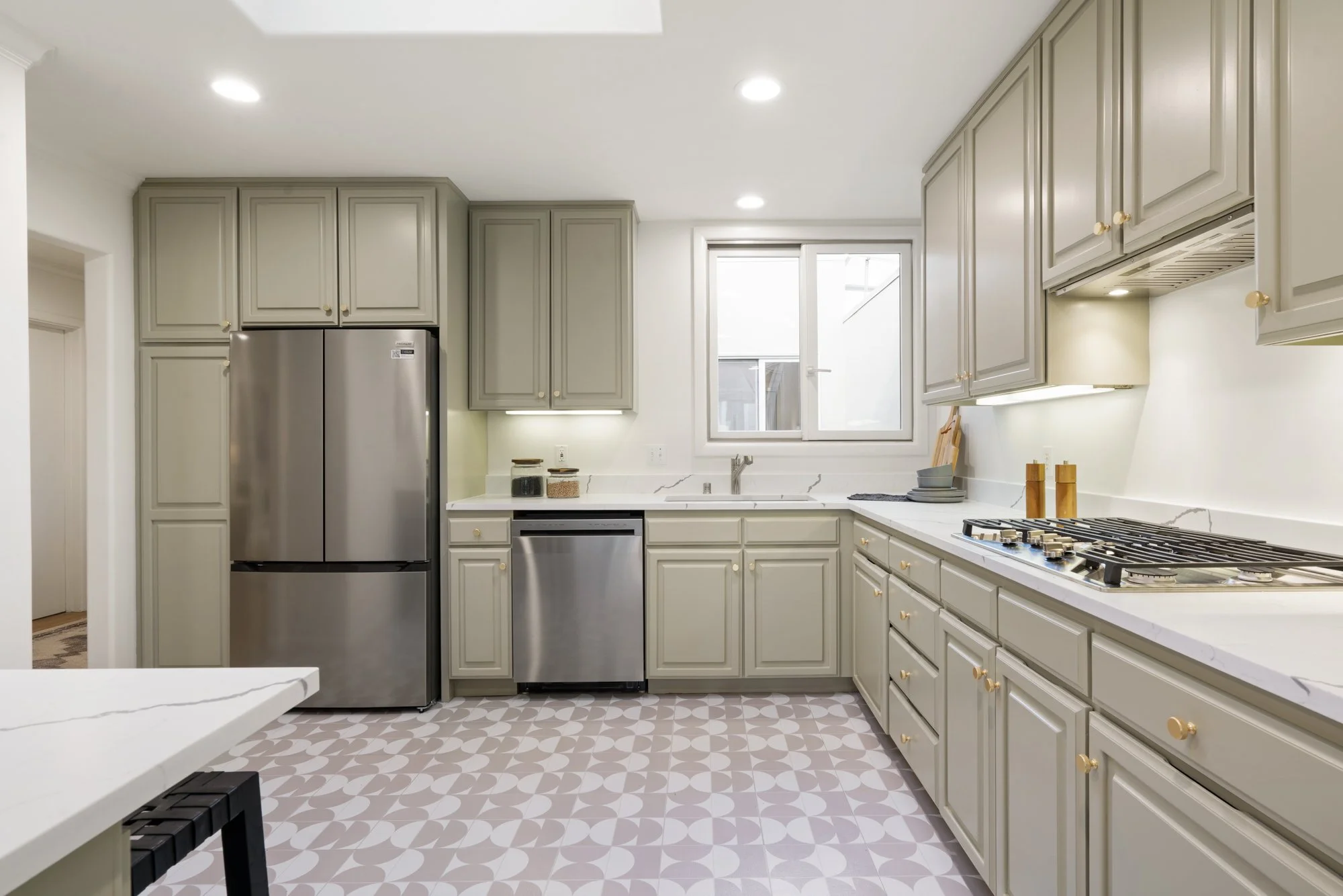 Modern kitchen with gray cabinets, stainless steel refrigerator and dishwasher, white marble countertops, patterned tile floor, bright lighting, window above the sink.