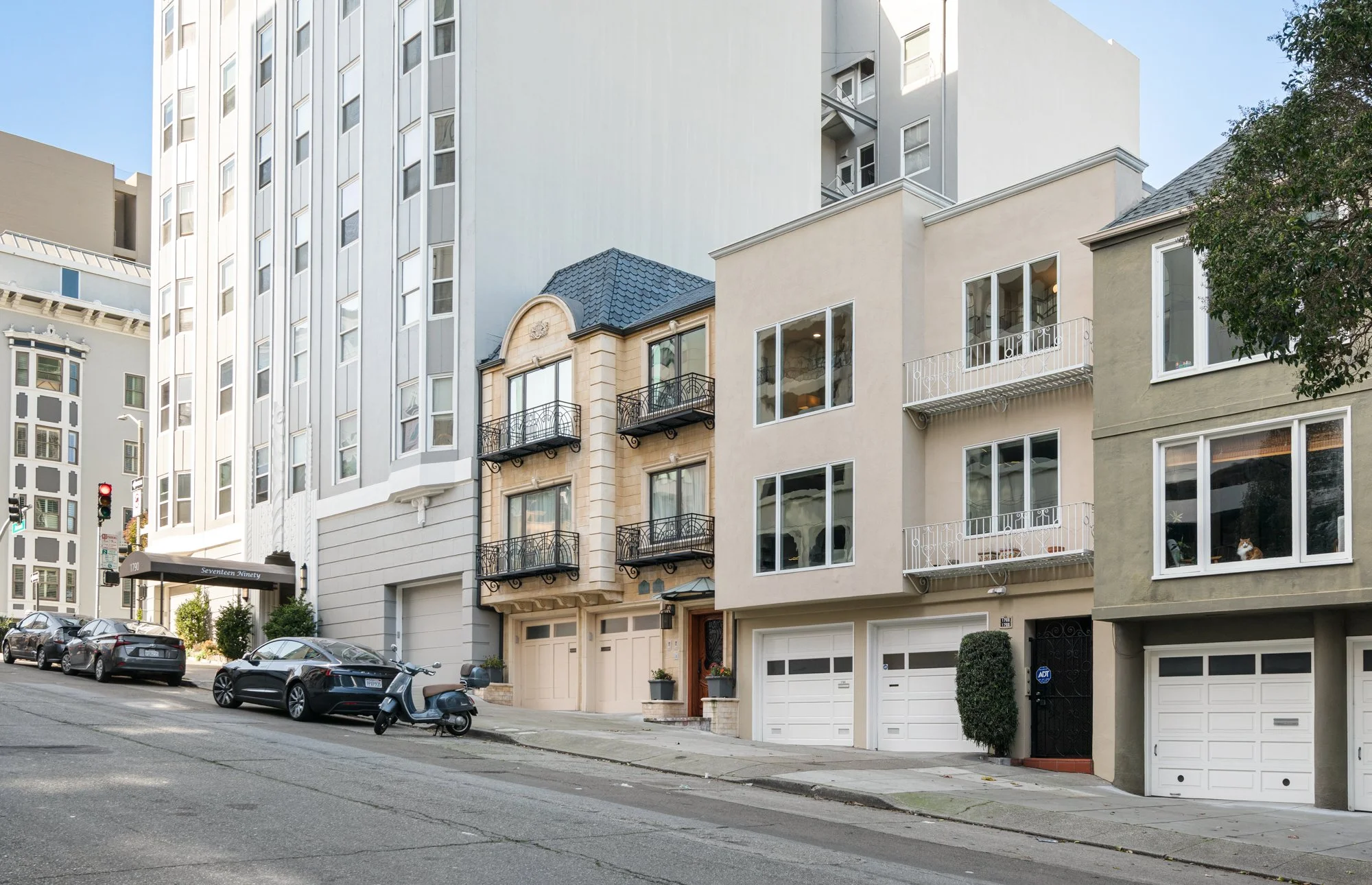 City street with parked cars, a scooter, modern residential buildings, and a hotel entrance in the background.