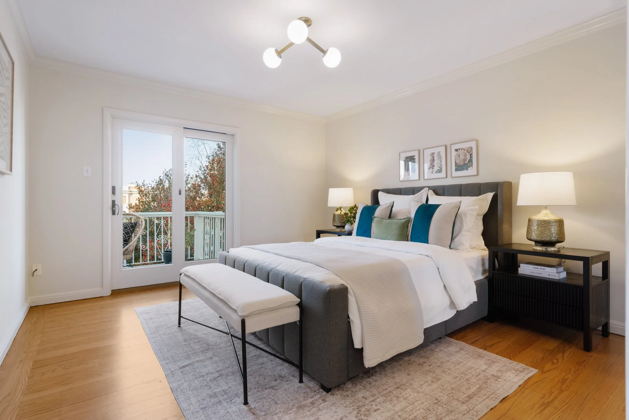 A modern bedroom with a large bed, gray upholstered headboard, white bedding with blue and green accent pillows, two black nightstands with table lamps, a bench at the foot of the bed, artwork on the wall, and a balcony door with a view of red and gr