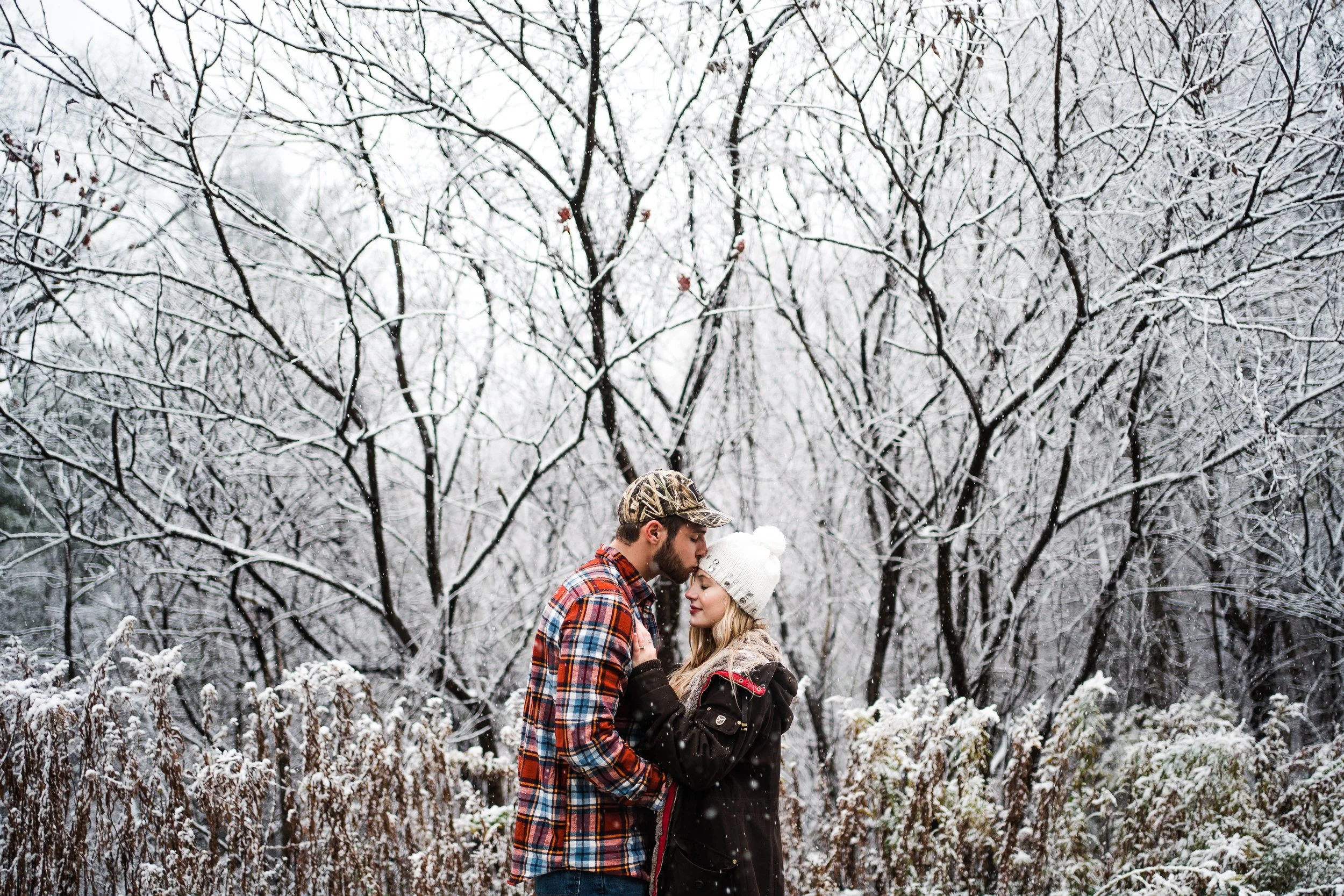 A couple stands close together in a snowy outdoor setting with snow-covered trees and bushes in the background, sharing an intimate moment.
