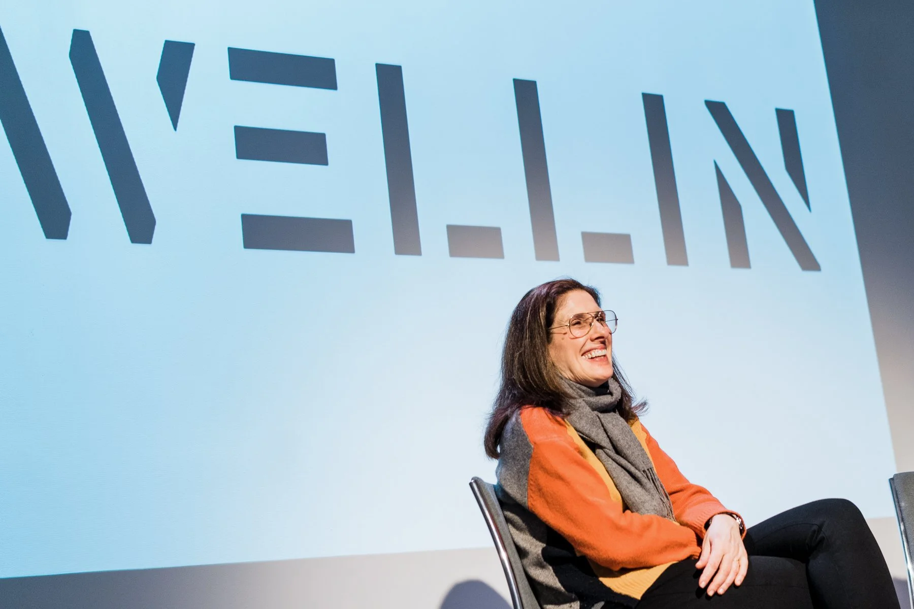 A woman sitting on a chair, smiling, wearing glasses and an orange and gray jacket, with a large screen behind her displaying the word 'WELLON' in bold letters.
