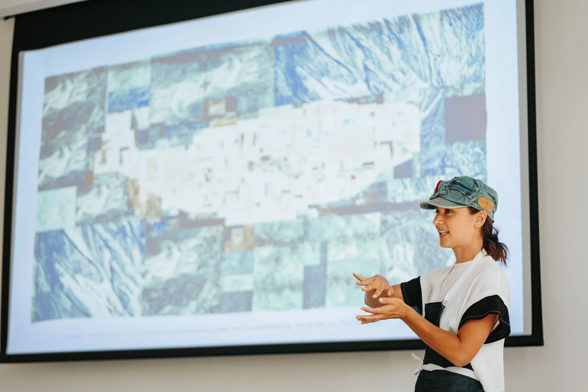Alexa Hatanaka giving a lecture on behalf of Wellin Museum. A woman wearing a denim cap and a white shirt is giving a presentation or explanation in front of a large screen displaying a detailed map or satellite image.
