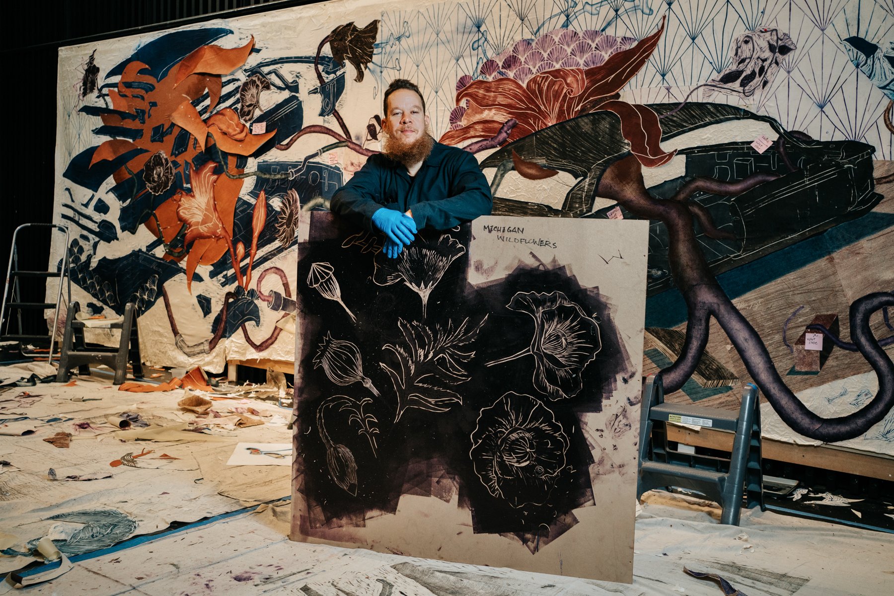 An artist with a beard and blue gloves standing behind a large floral artwork on an easel in a workspace. The background features a colorful mural of flowers, vines, and abstract designs on a wall. The floor has scattered papers and art supplies, and
