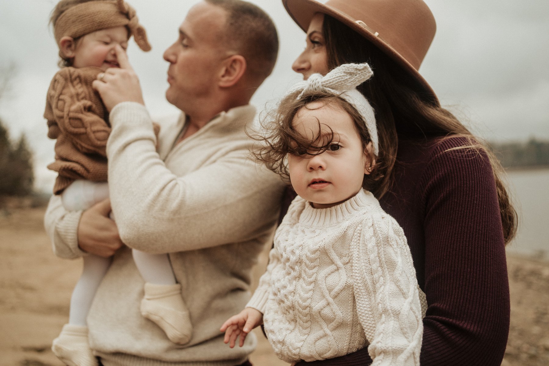 A family of four outdoors with two women, one man, and two young girls. One woman is wearing a wide-brimmed hat and is close to one girl, who is wearing a cardigan and headband. The man is holding the other girl, who is wearing a sweater and sitting 