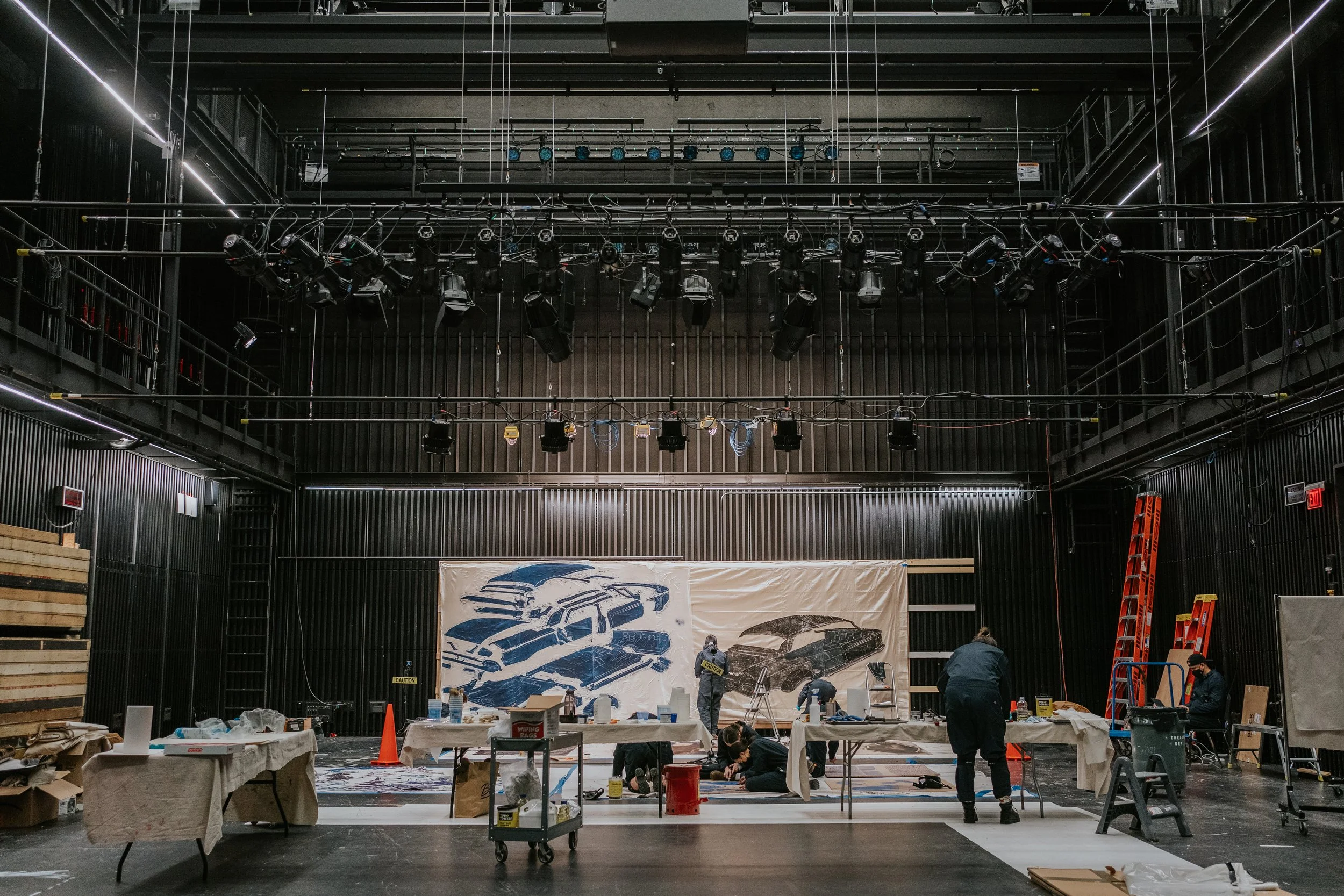 Collaboration with Yashua Klos and Wellin Museum. People setting up and painting a large automotive-themed mural inside a modern industrial-style venue with black walls and ceiling, and stage lighting.
