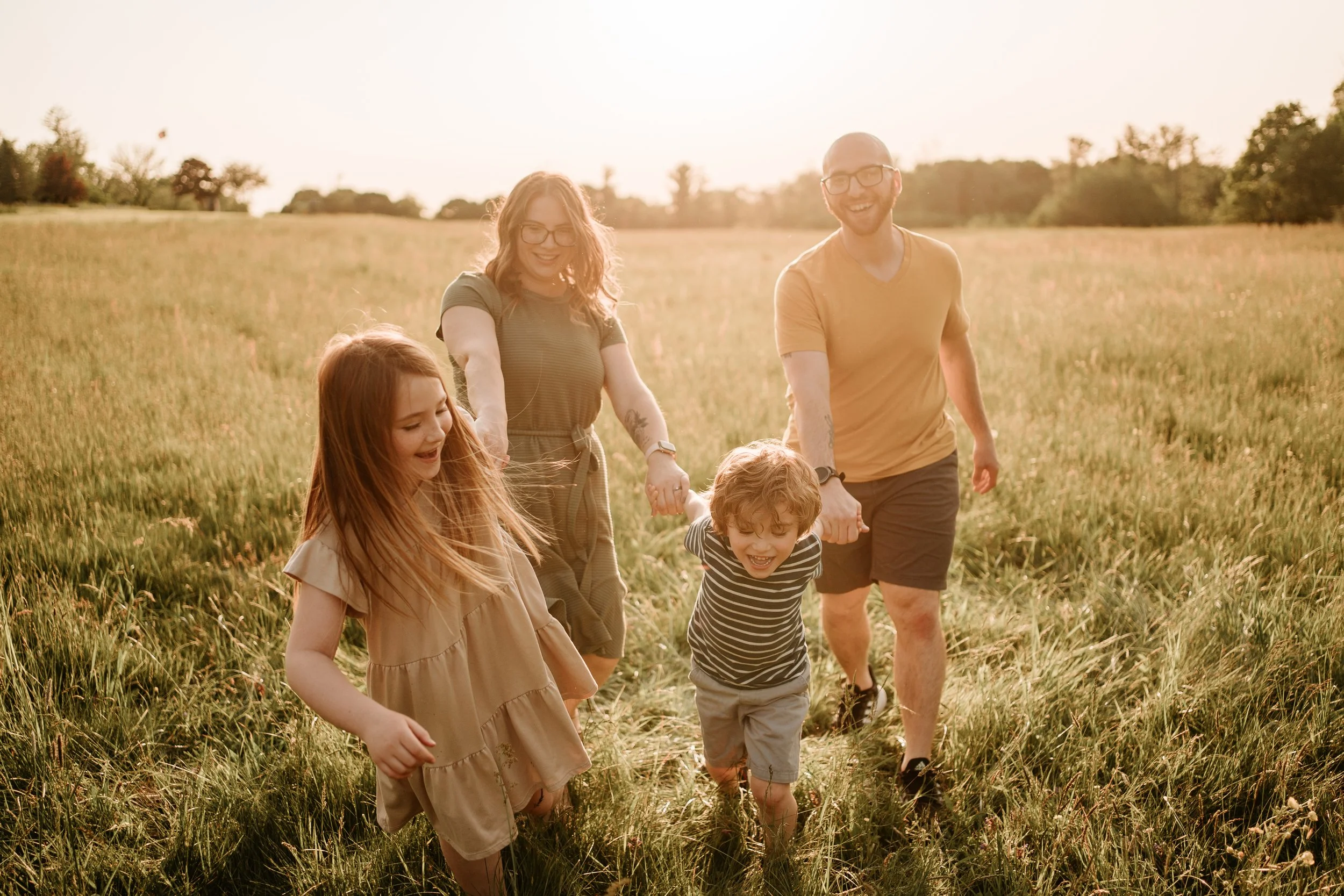 A family of five, including two adults and three children, holding hands and walking together through a grassy field during sunset, smiling and enjoying the outdoors.