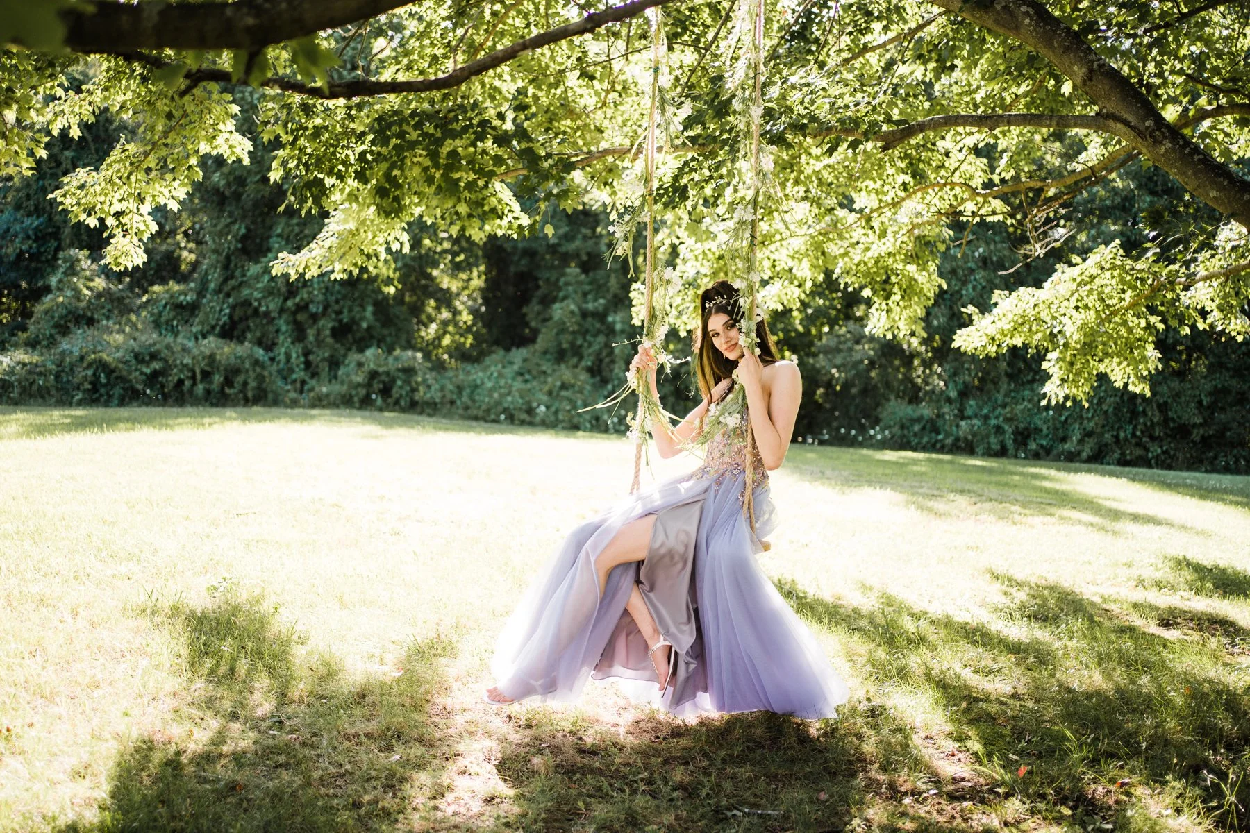 A woman in a flowing lavender gown sitting on a swing hanging from a tree in a sunlit grassy area surrounded by green trees.