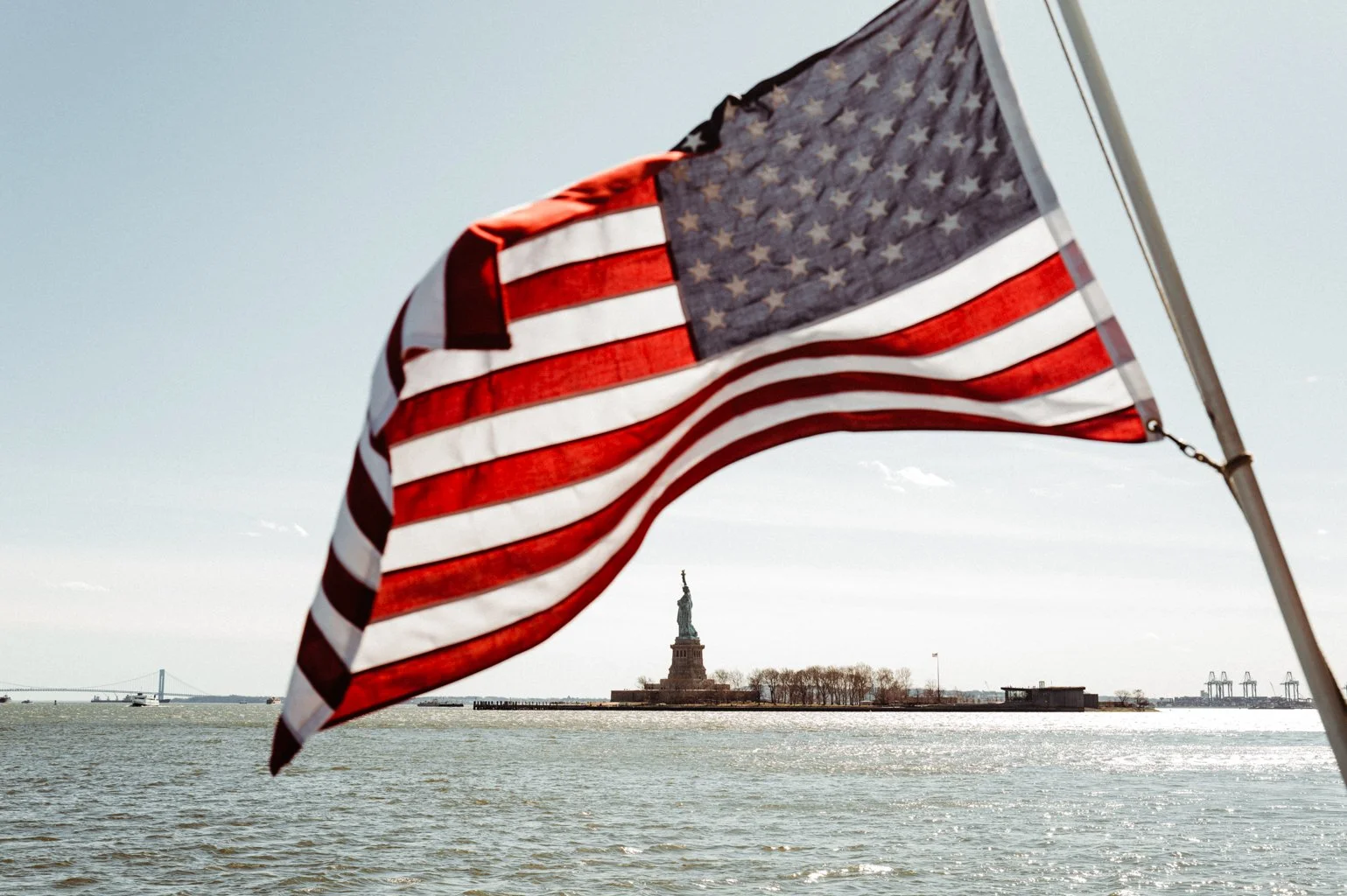 American flag in the foreground with the Statue of Liberty in New York Harbor in the background.