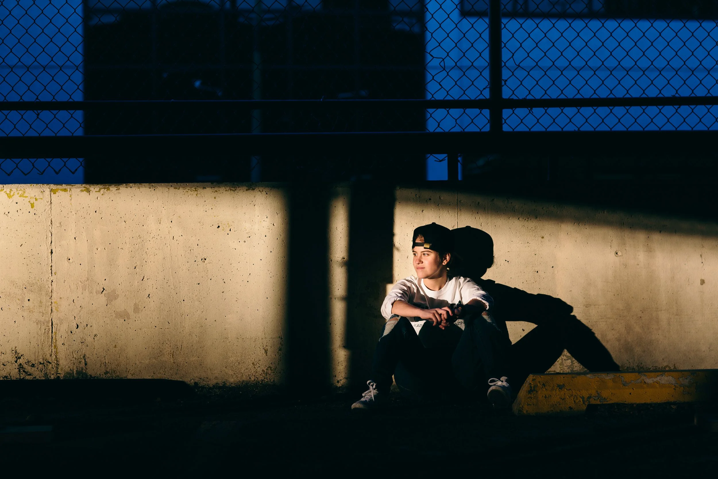 A young woman sits against a concrete wall in sunlight, wearing a black baseball cap backwards, a beige jacket, and black shoes. She appears to be relaxed, with crossed arms, in an outdoor setting with a chain-link fence overhead.