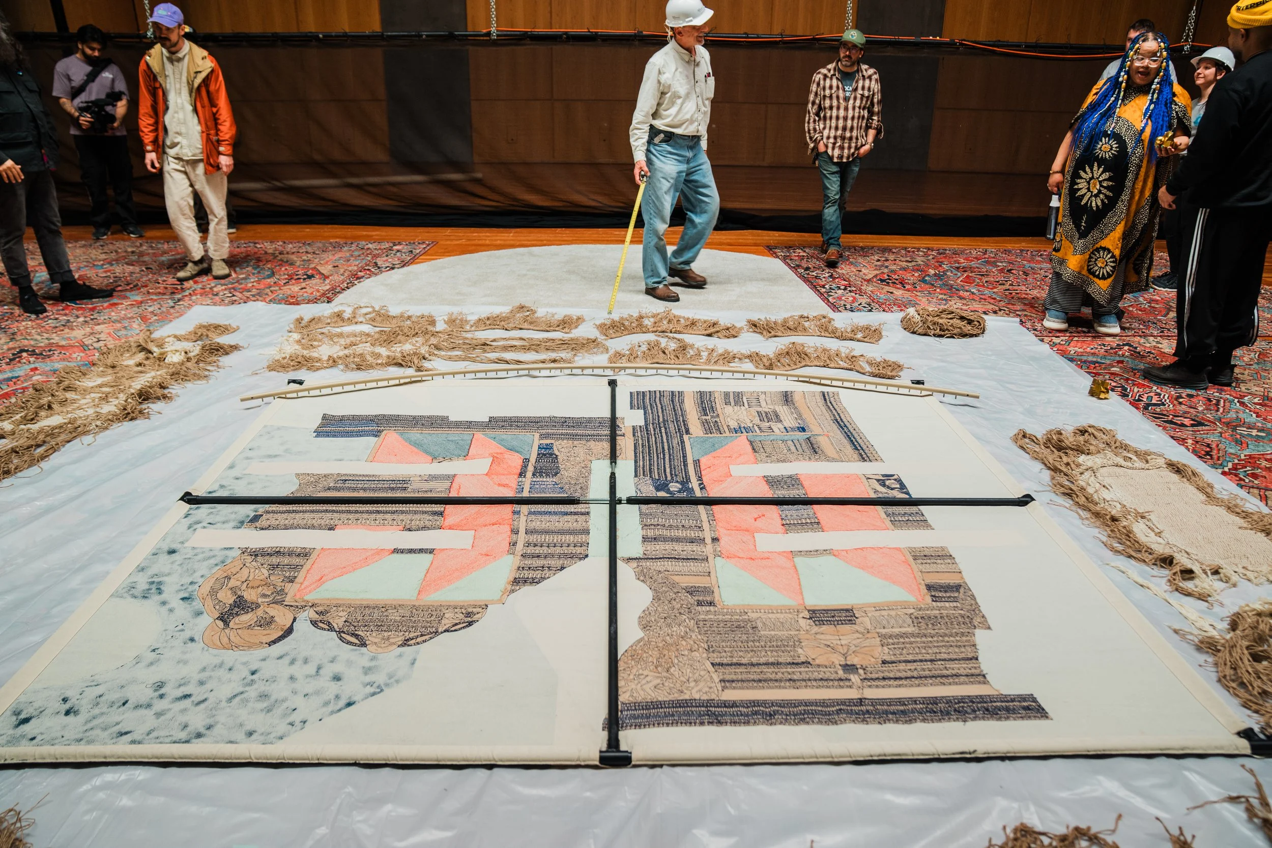 Group of people, some wearing hard hats, standing around a large object or installation with textile and cardboard materials, in an indoor setting with wooden walls.