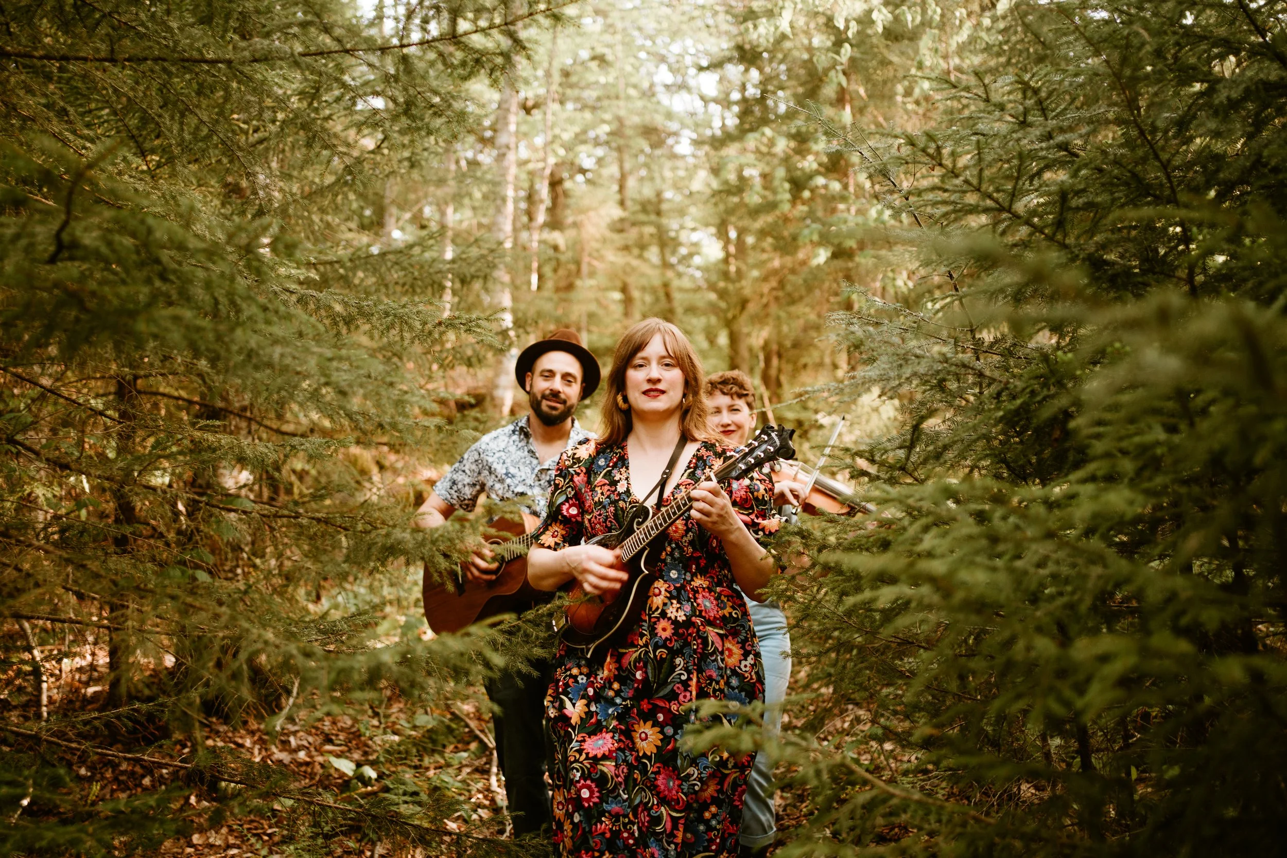 Three people walking in a forest, with a woman in the front playing a guitar, and two others behind her, one with a ukulele and another with a violin, among green trees.