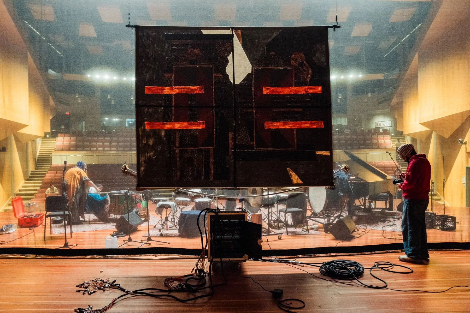 The image shows an auditorium stage with a large black and red abstract art piece hanging in the center. There are two men working on the stage: one on the right wearing a red hoodie and checkered hat, using a camera, and another on the left in a tan jacket and blue head covering, sitting near some equipment. The stage is set with various musical instruments and equipment, and there are stairs on both sides leading to the upper level. The auditorium has wooden walls and tiered seating visible in the background.