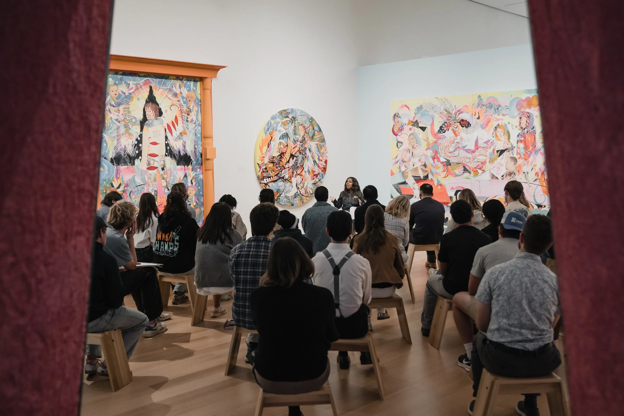 A lecture by Jamea Richmond-Edwards. An art gallery with colorful abstract paintings on the walls. A woman is speaking to an audience of diverse people seated on benches, listening attentively.