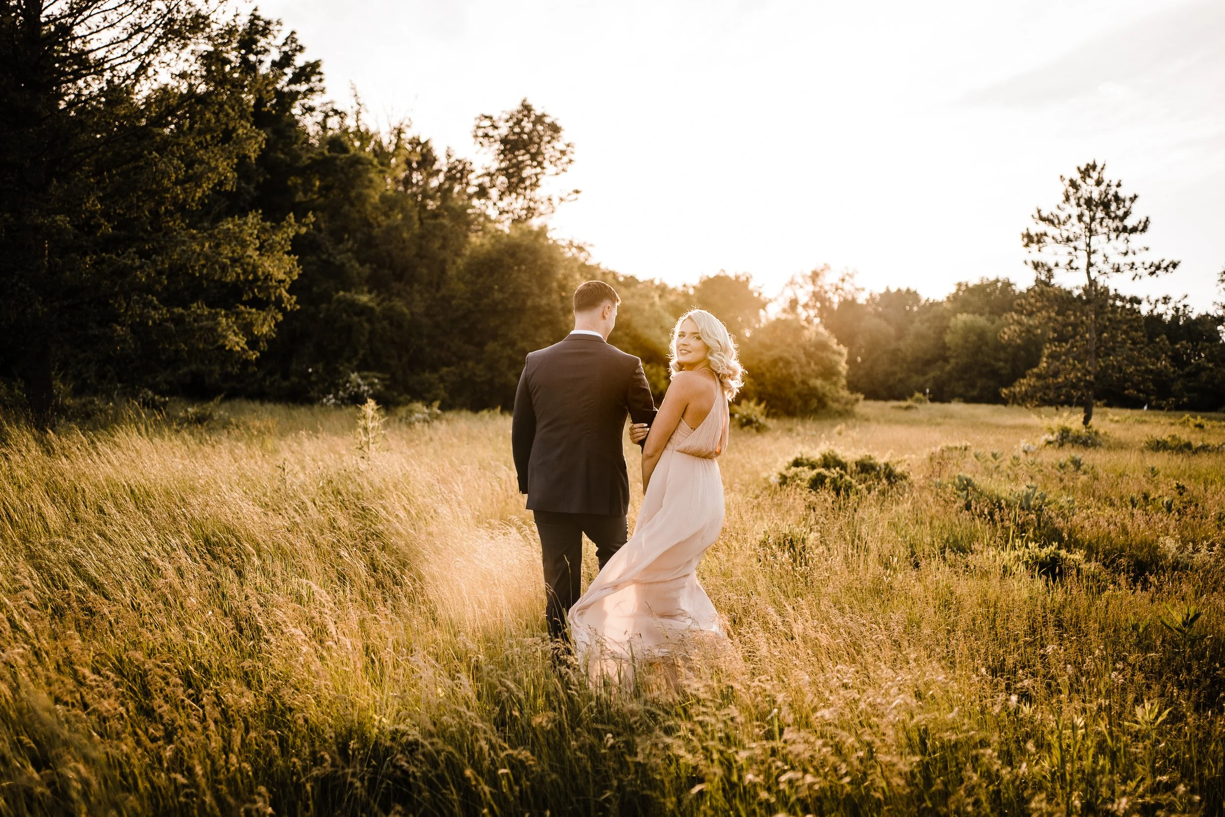 A couple standing in a grassy field during sunset, facing away from the camera. The woman is wearing a flowing dress and the man is in a suit. They are holding hands and looking at each other or the scenery.