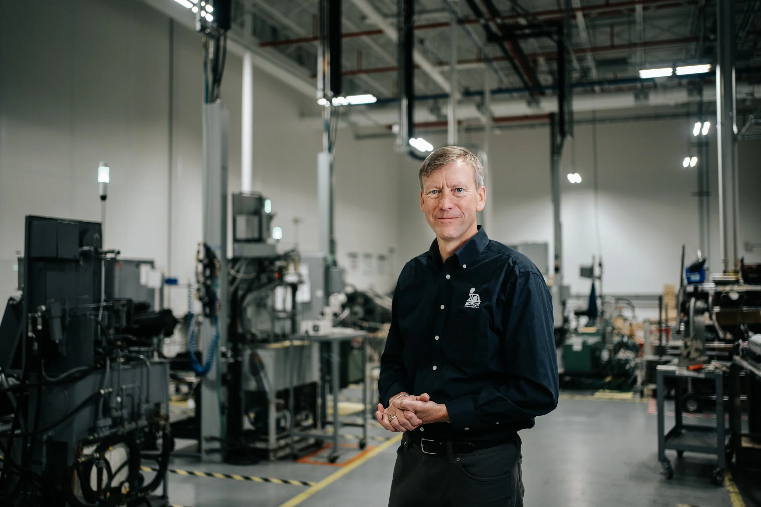A man in a black shirt standing inside a manufacturing or industrial facility with machinery and equipment in the background.