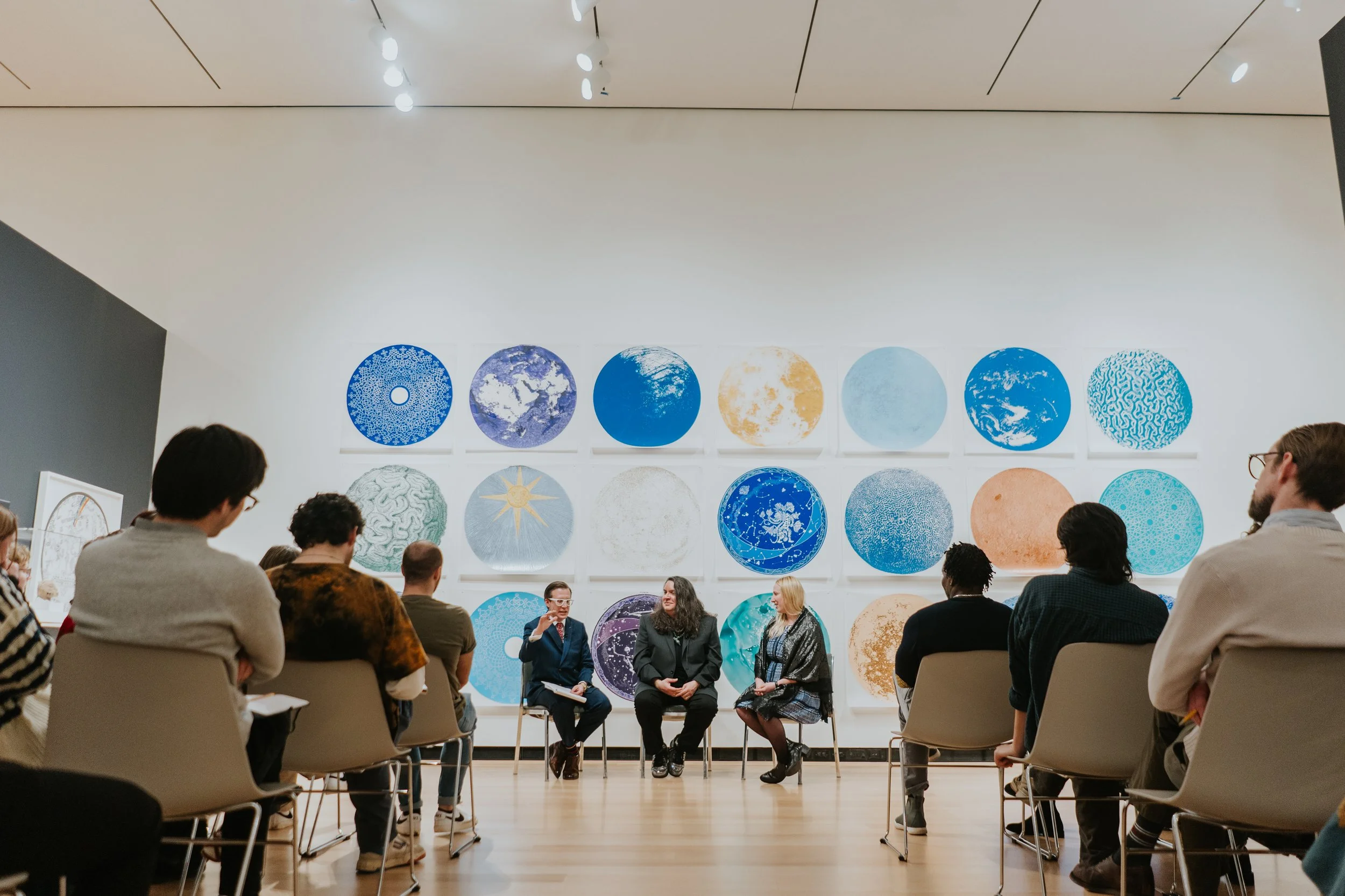 Group of people sitting and standing in a room listening to a panel of three women and a man engaged in discussion, with a large wall of circular artwork behind them.