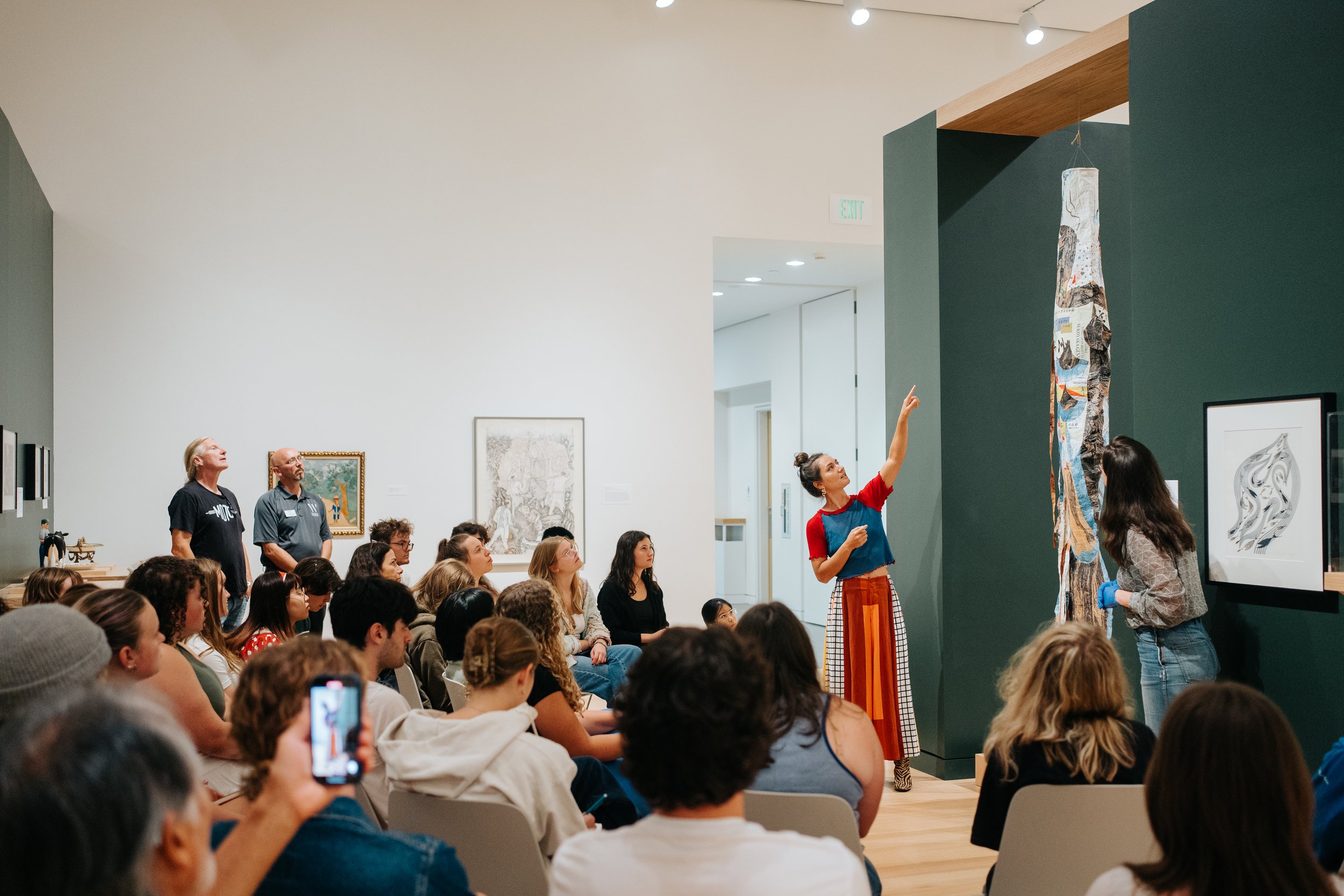 A lecture by Alexa Hatanaka. A woman in colorful clothing is gesturing towards a tall art display in front of a seated audience inside an art gallery with various framed artworks on the white and dark green walls.