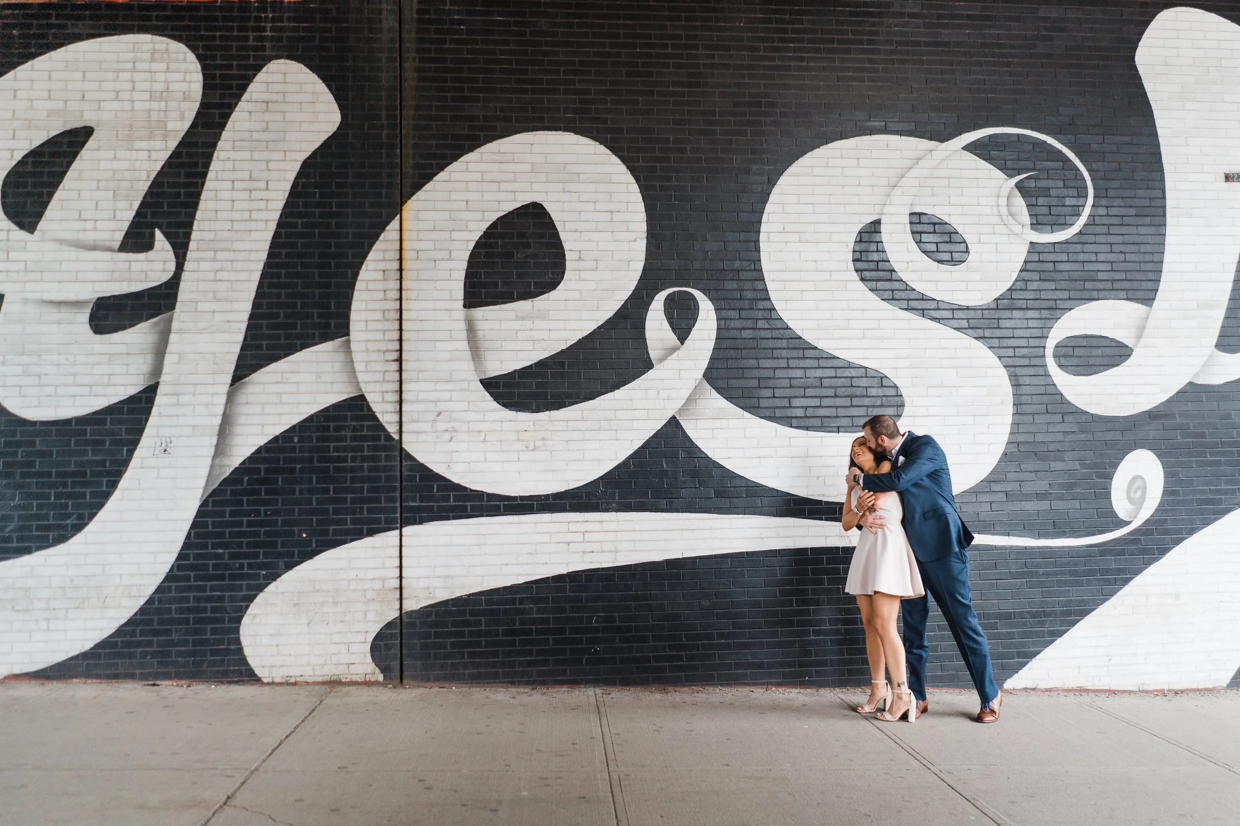 A couple dressed in formal attire sharing a kiss in front of a large black and white mural on a brick wall.