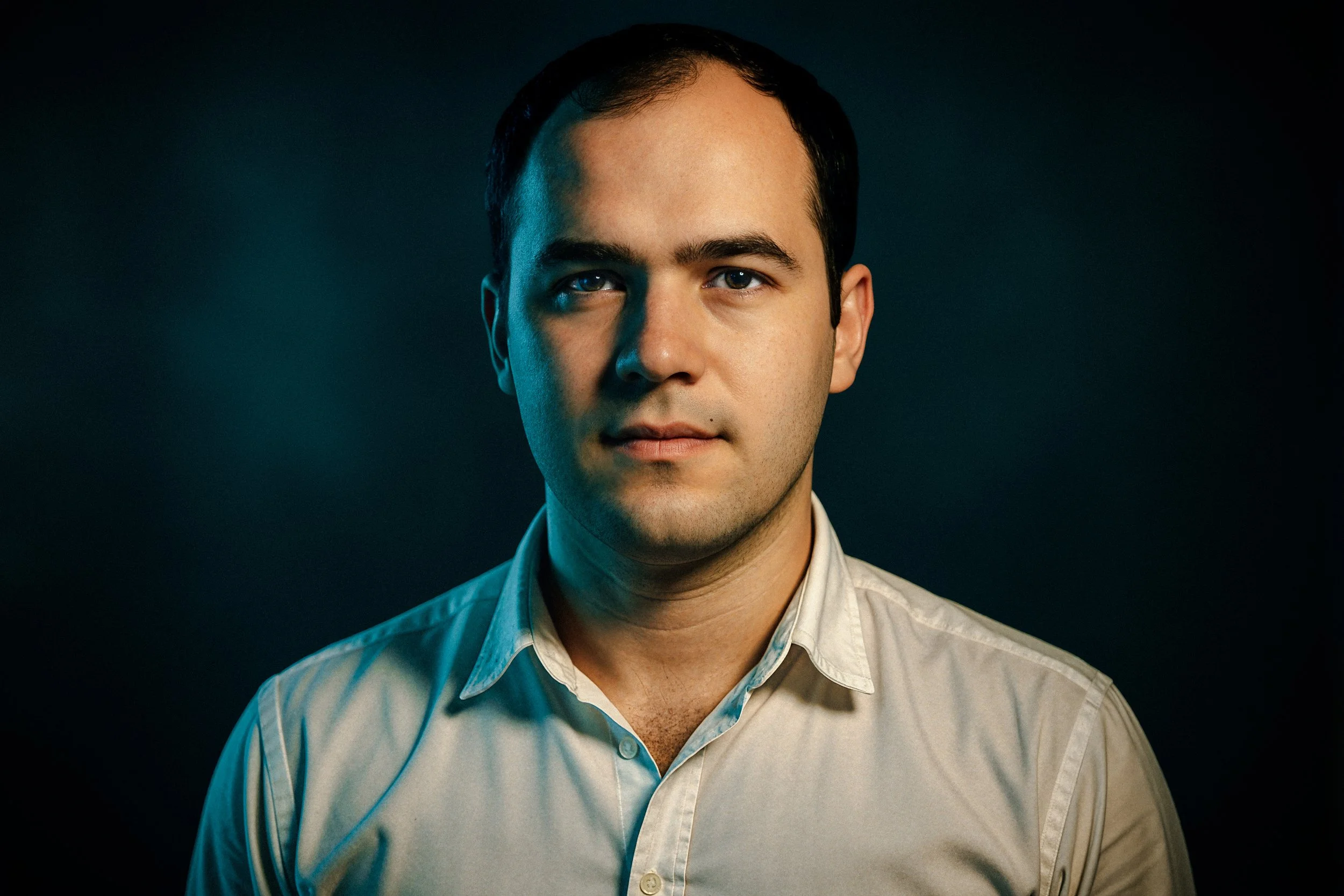 Portrait of a man with short dark hair, wearing a light-colored dress shirt, against a dark background.