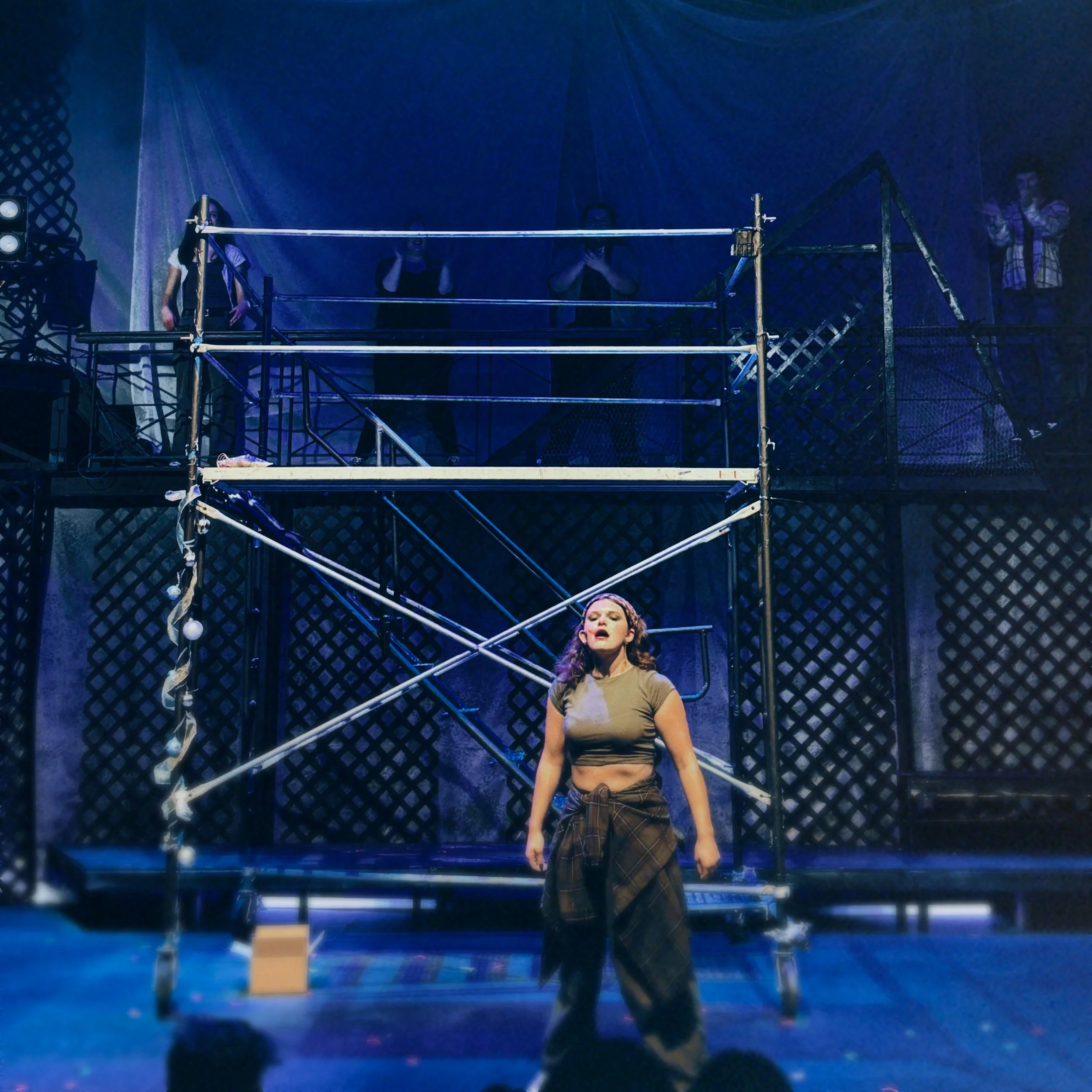 A young woman sings on a dimly lit stage, standing in front of scaffolding and set pieces.