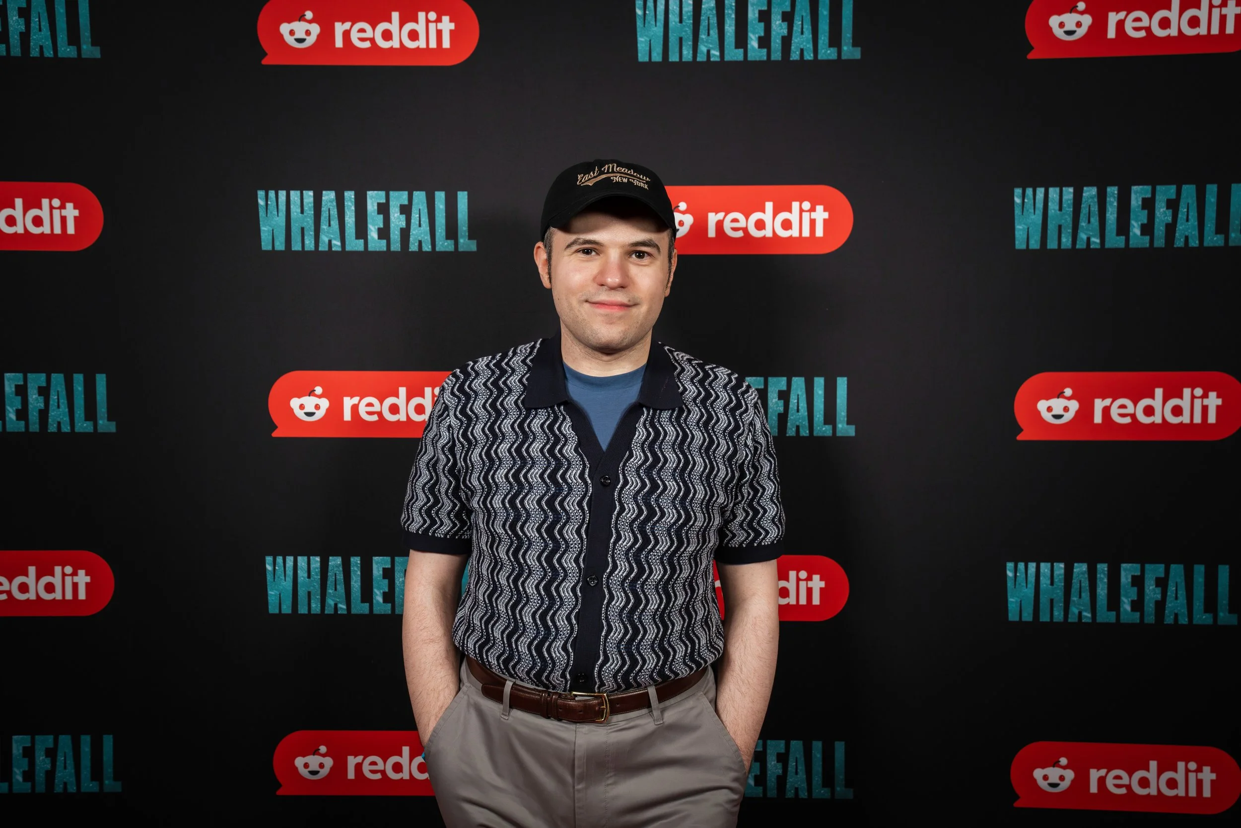 Man in a black cap, patterned shirt, and gray trousers poses with a slight smile against a black Whalefall and Reddit event backdrop.