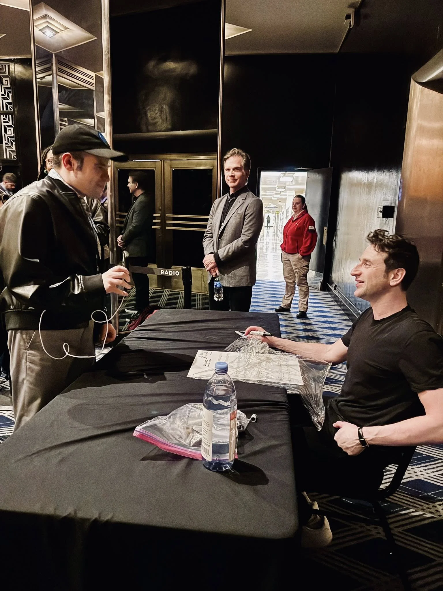A man in a black t-shirt sits at a table, smiling up at another man in a cap and leather jacket. They are at a formal venue with patterned flooring. Several people stand in the background.