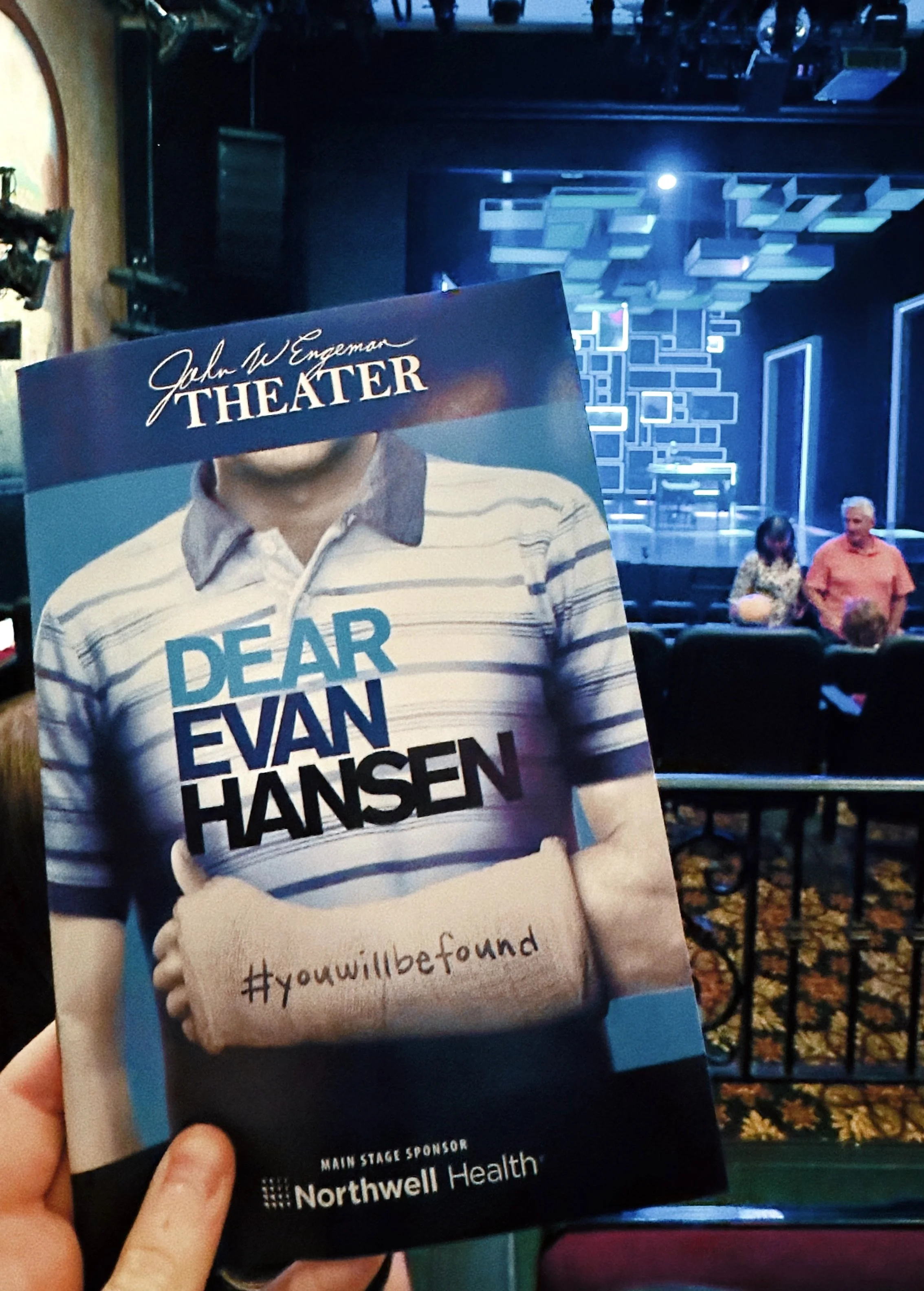 Hand holding a Dear Evan Hansen theater program in front of a blue-lit stage.
