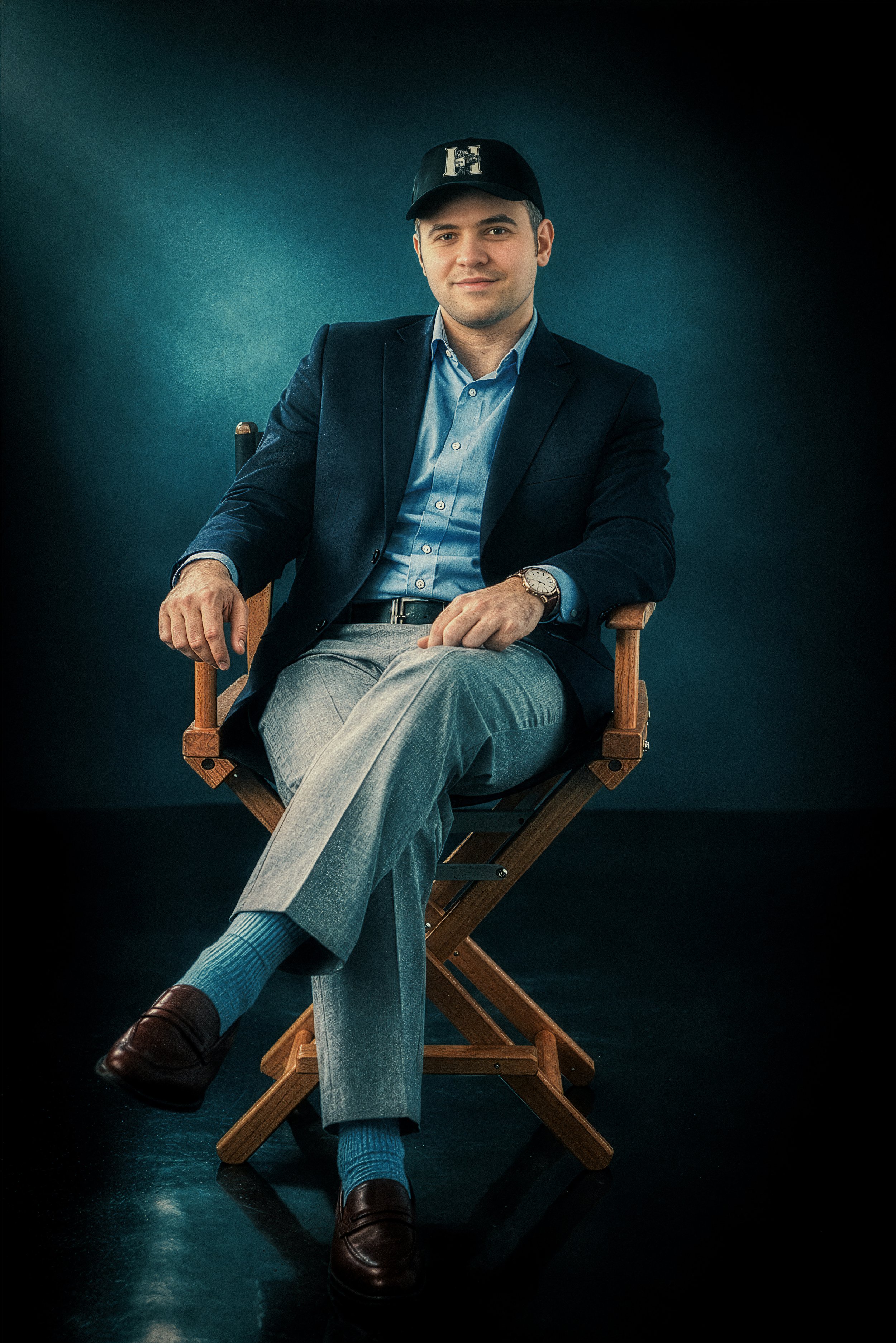 A young man sitting on a director's chair, wearing a navy blazer, light blue shirt, grey pants, blue socks, and brown shoes, with a black baseball cap and a wristwatch, against a dark background.