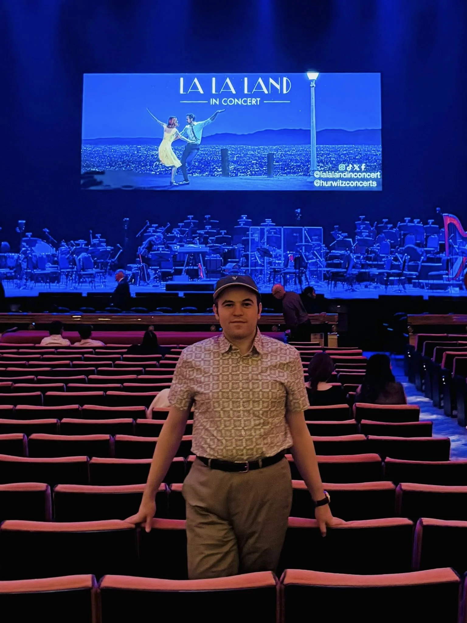 A person stands in a theater in front of an empty stage. A "La La Land in Concert" poster is projected onstage.